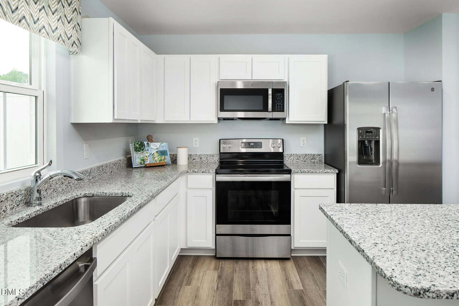 Modern kitchen with white cabinets, stainless steel appliances including a refrigerator, oven, and microwave, granite countertops, a double sink, and light blue walls. Wood-look flooring is visible.