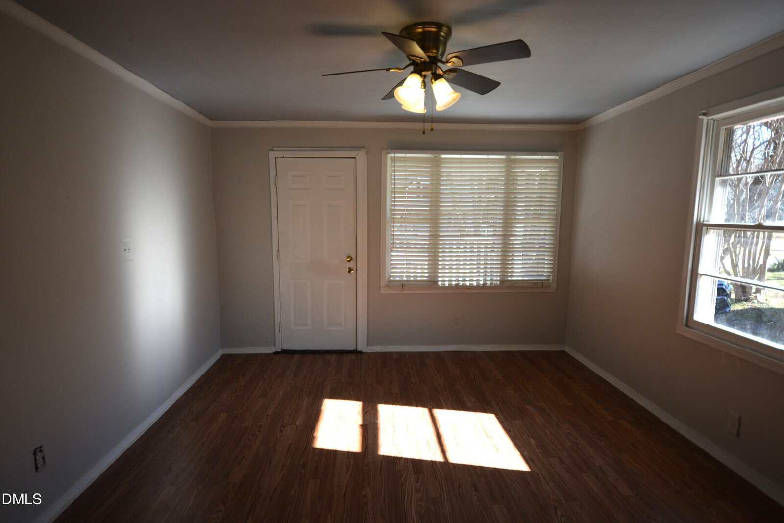 A small empty room with light brown wood flooring, beige walls, a white front door, a window with blinds, a side window, and a ceiling fan with lights turned on. Sunlight is shining through the windows onto the floor.