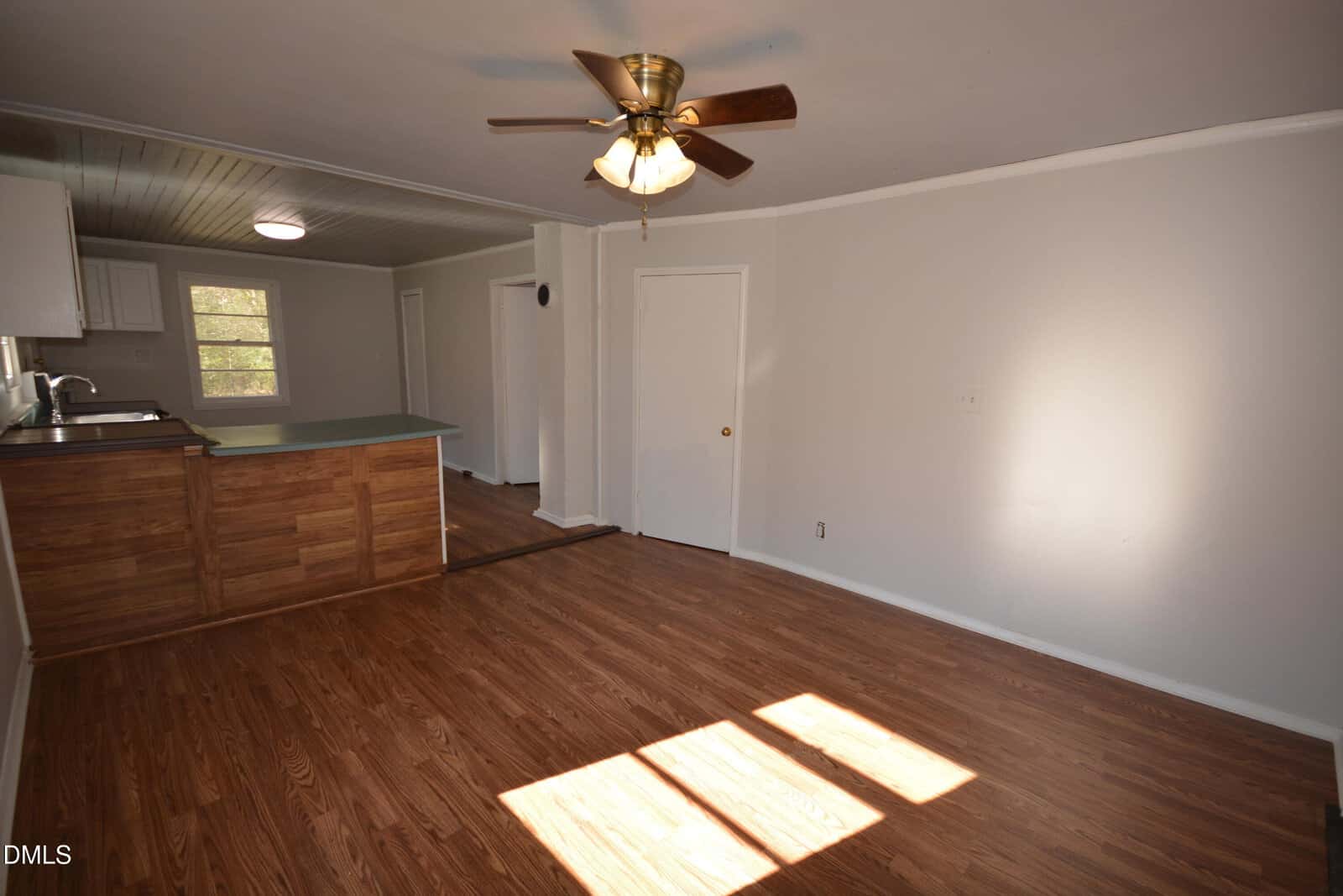 A mostly empty room with wood laminate flooring, white walls, a ceiling fan with lights, and a partial view of a kitchen area with a wood-paneled counter and white cabinets. Sunlight enters through a window.