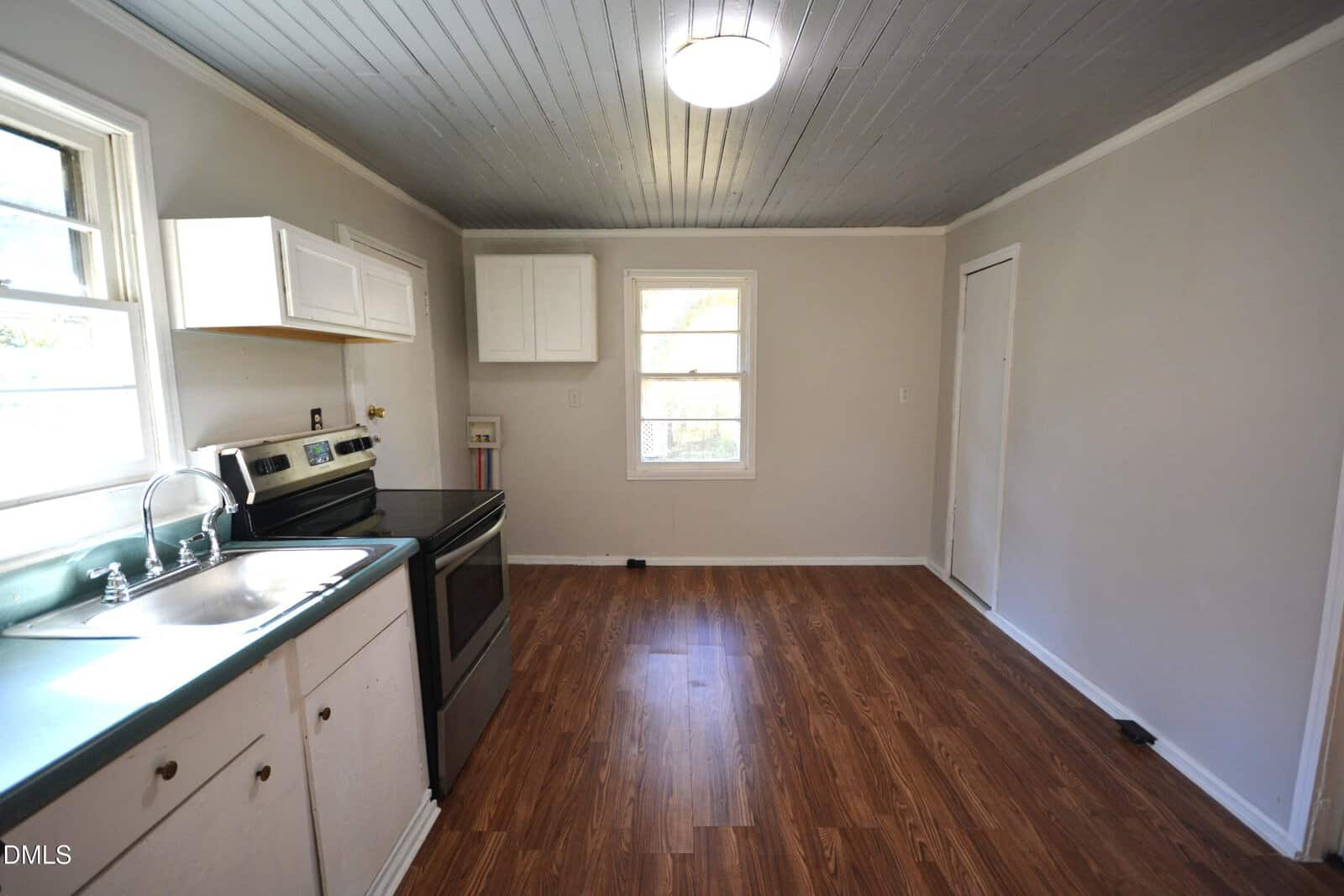 A small kitchen with wood-look flooring, white cabinets, a stainless steel stove, a stainless steel sink under a window, and light gray walls with a gray plank ceiling. A single ceiling light is on.