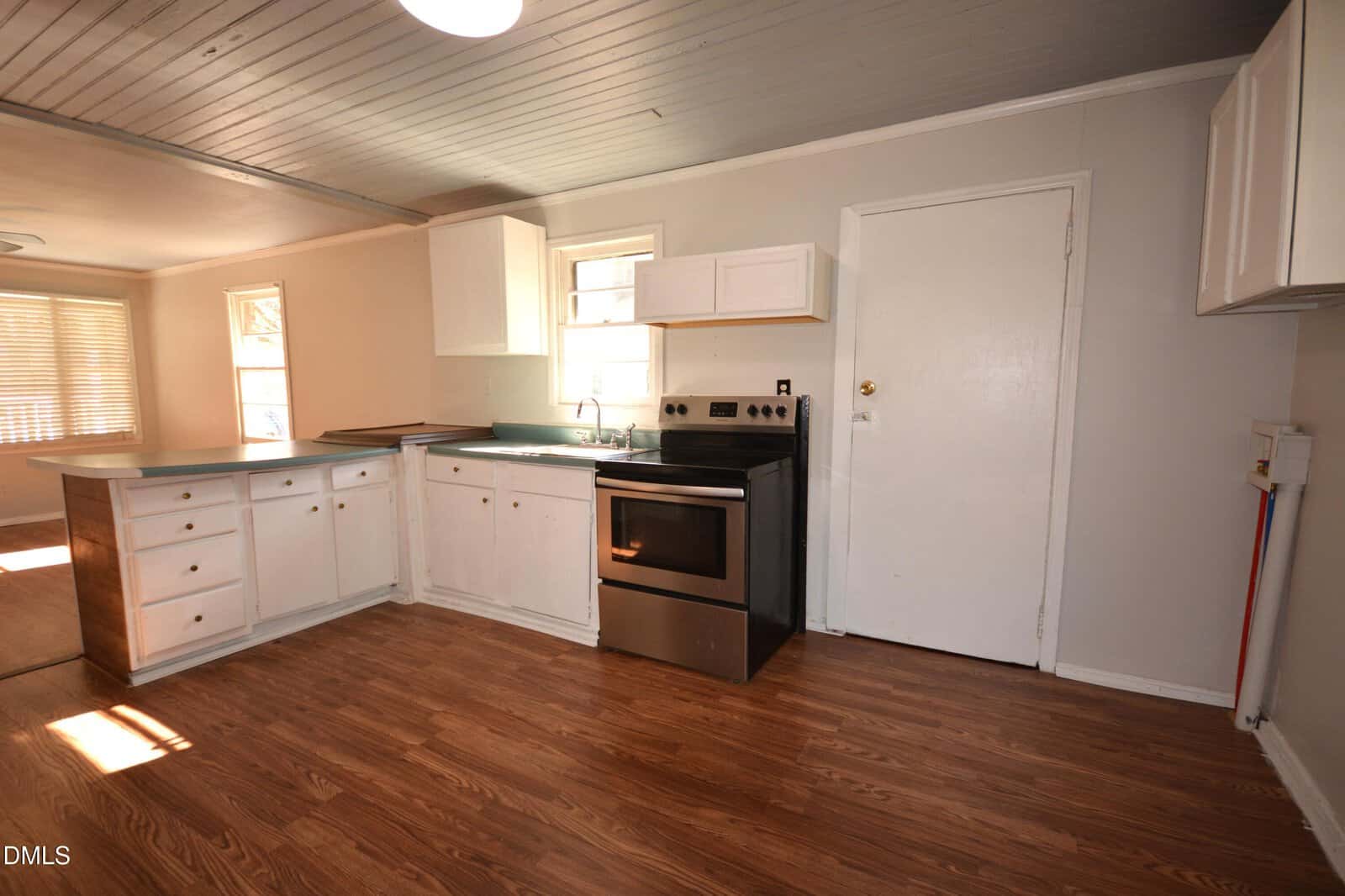 A kitchen with white cabinets, a stainless steel electric stove, a sink under a window, wood-patterned flooring, light-colored walls, and a ceiling with visible paneling. There is a door and open countertop space.