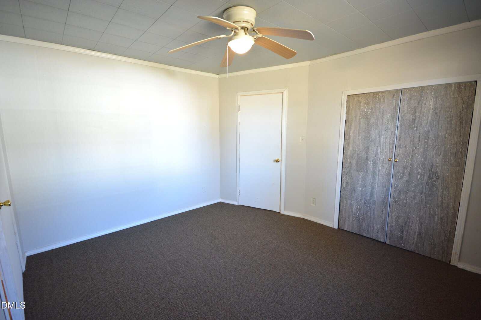 Empty room with white walls, brown carpet, a ceiling fan with light, a closed white door, and a closet with wood-patterned sliding doors. The room appears clean and unfurnished.