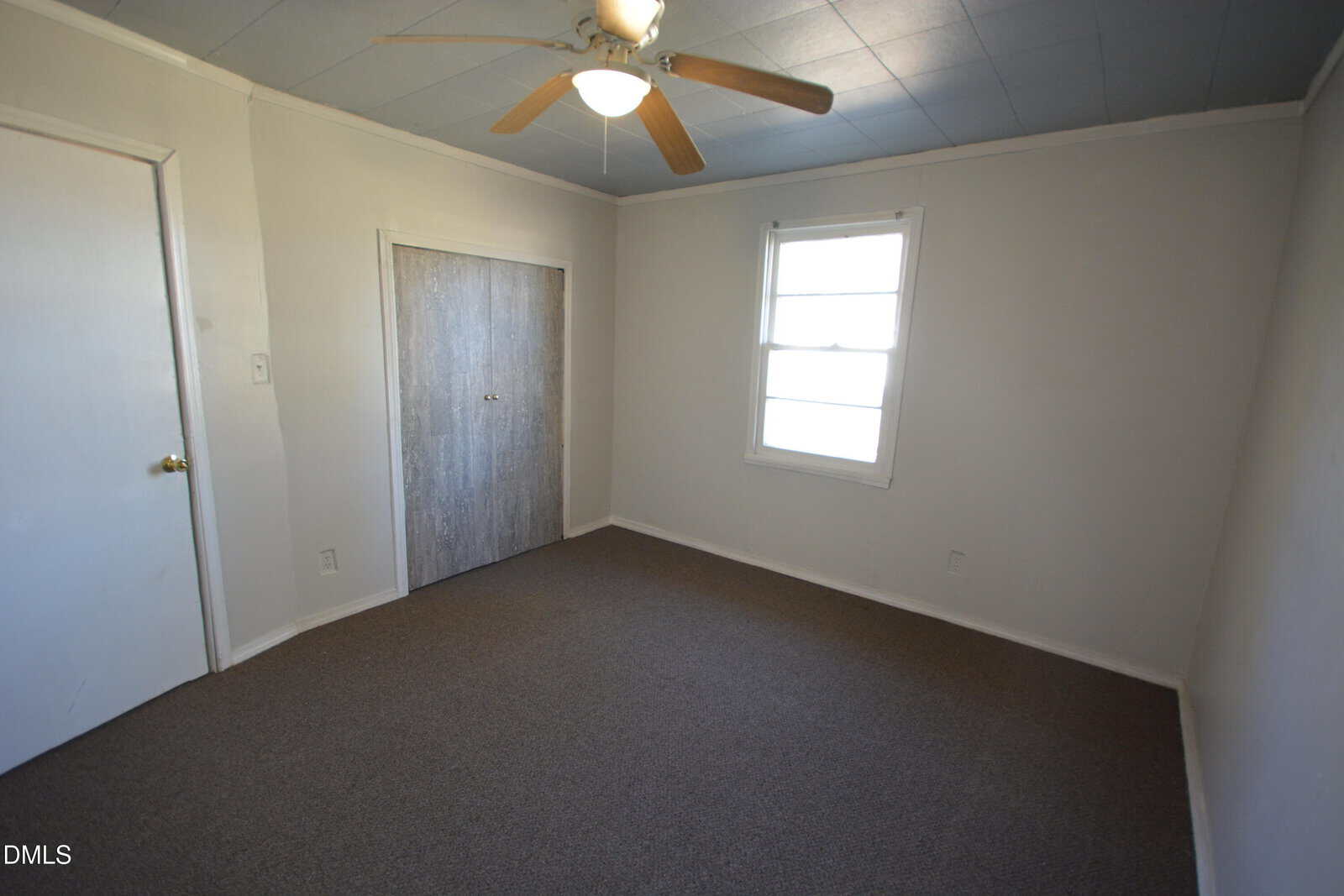 Small, empty room with beige carpet, white walls, a window, closed closet with wooden doors, ceiling fan with light, and a closed door on the left wall.