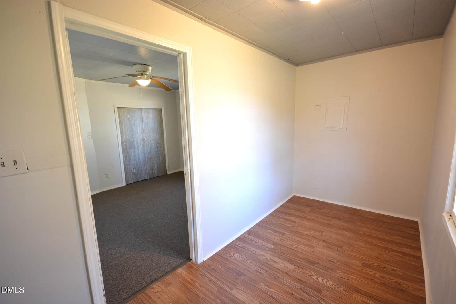 A small, empty room with wood flooring and white walls connects to a carpeted room with a ceiling fan and closed wooden doors. A window lets in natural light on the right wall.