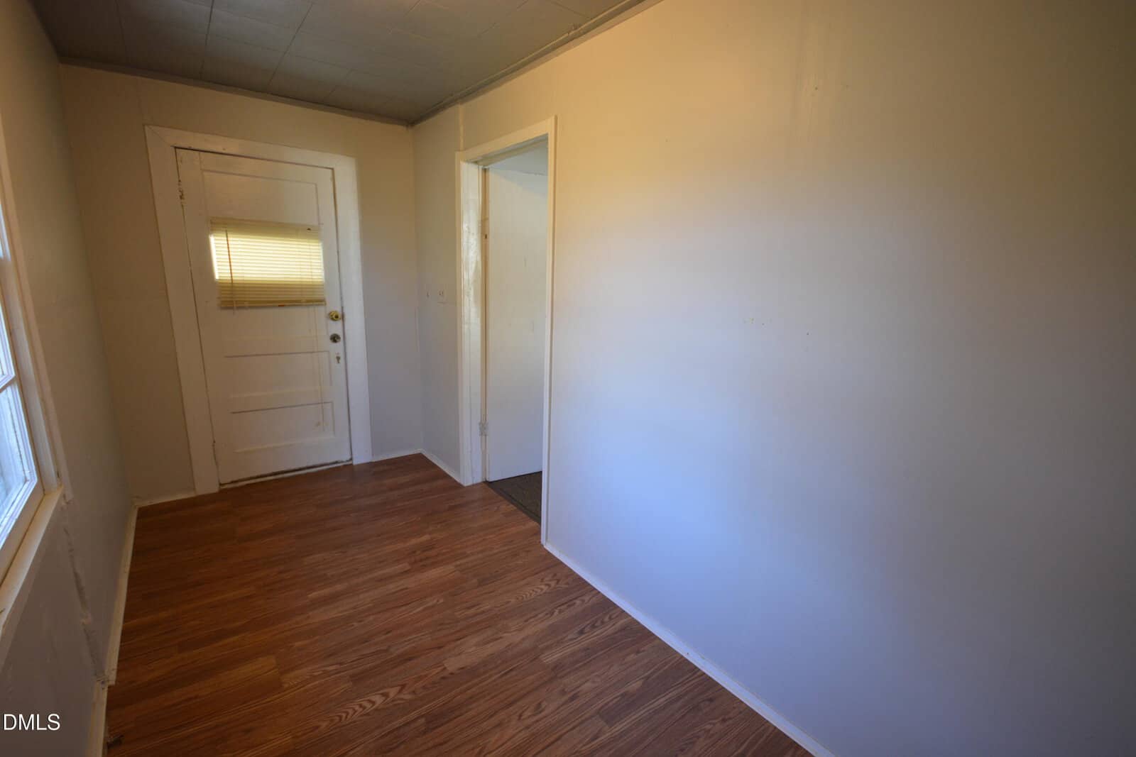 Small entryway with wood-style flooring, white walls, a white door with a window and blinds, and a window on the left. The hallway leads to another room through a large open doorway.