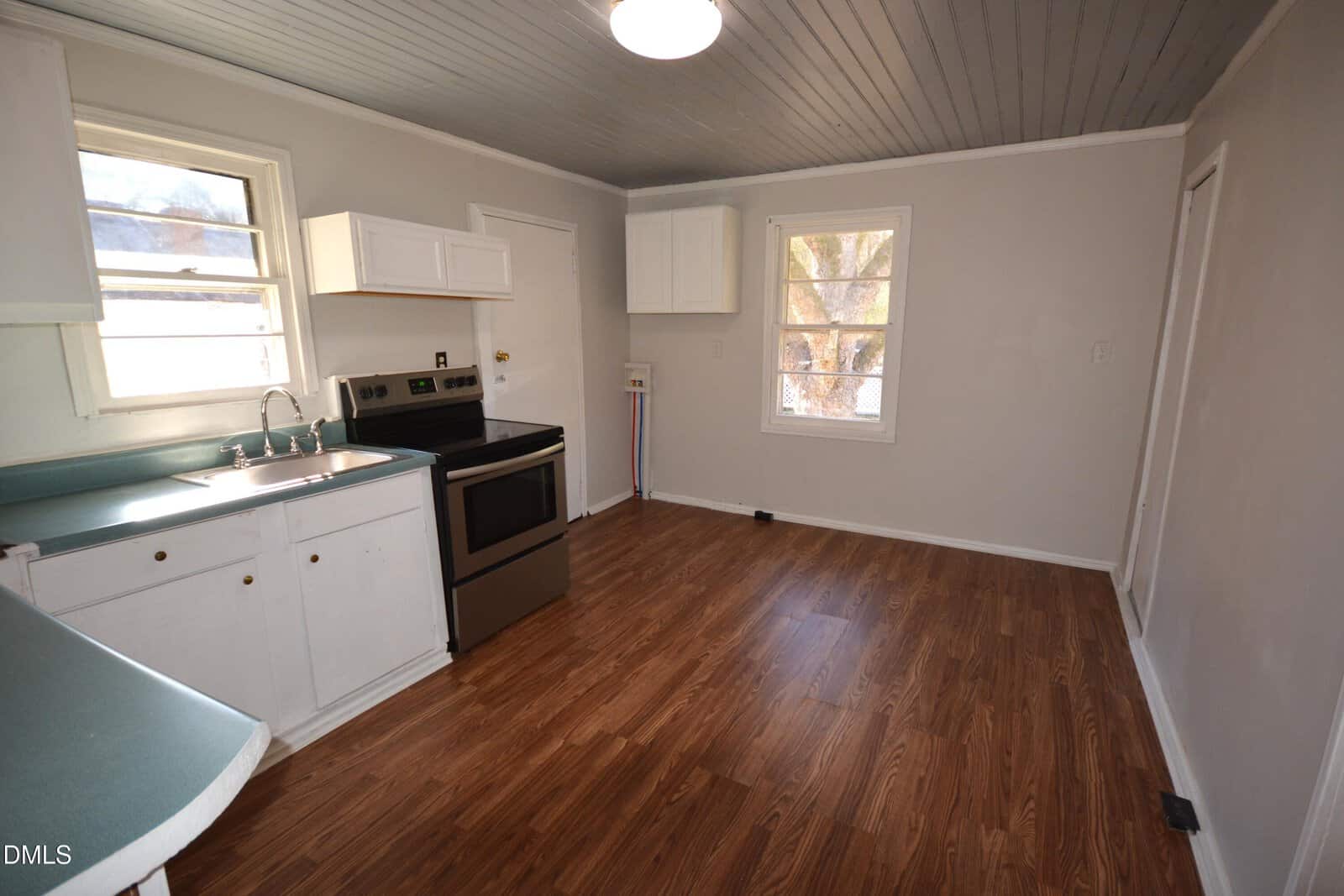 A small kitchen with white cabinets, a stainless steel stove, single sink, green countertops, and wood laminate flooring. There are two windows and a ceiling light. The walls are painted light gray.