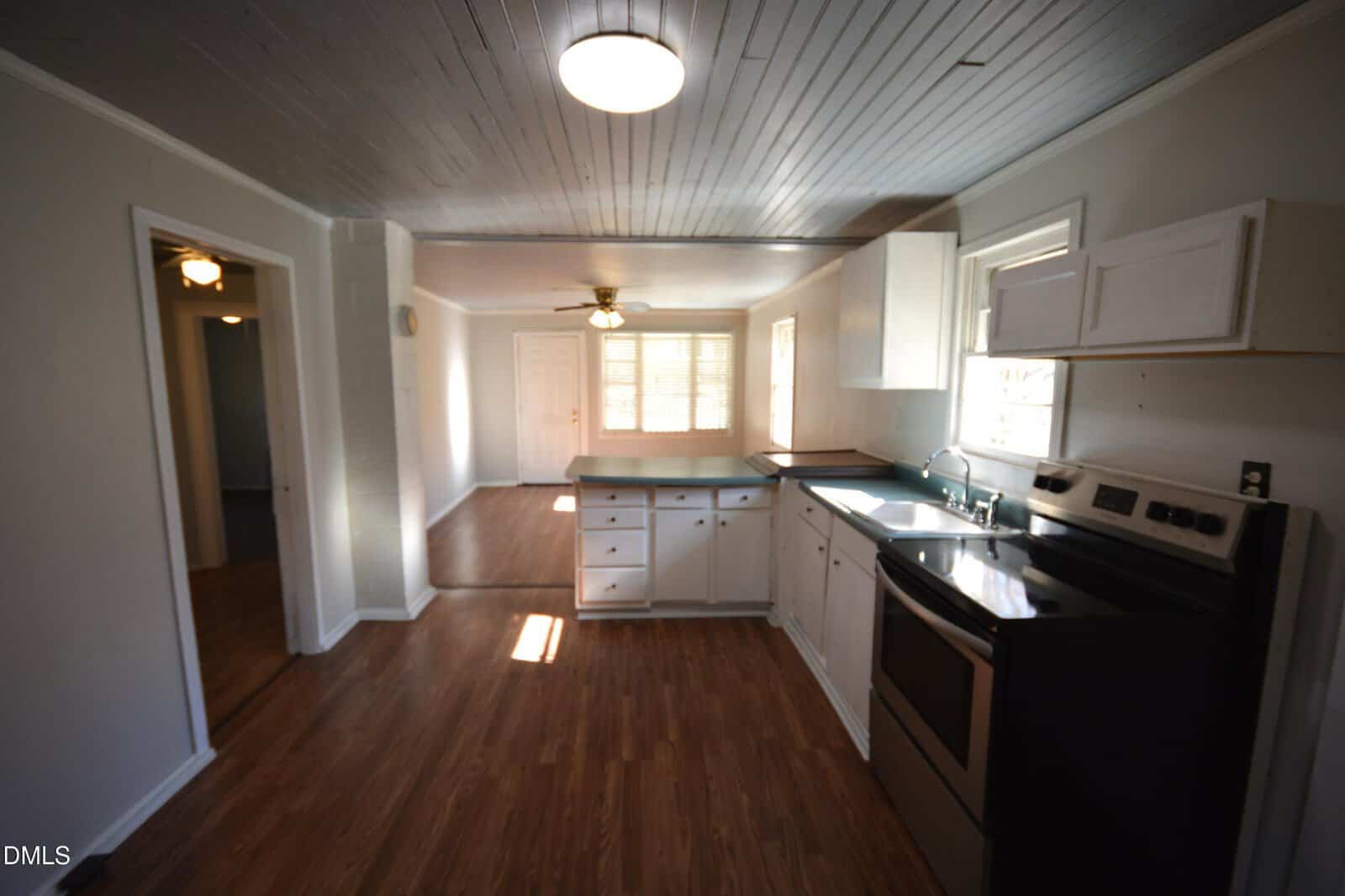 A kitchen with white cabinets, stainless steel appliances, and a double sink, opening into a dining area with wood-style floors, gray walls, and a ceiling fan. Natural light comes in through several windows.