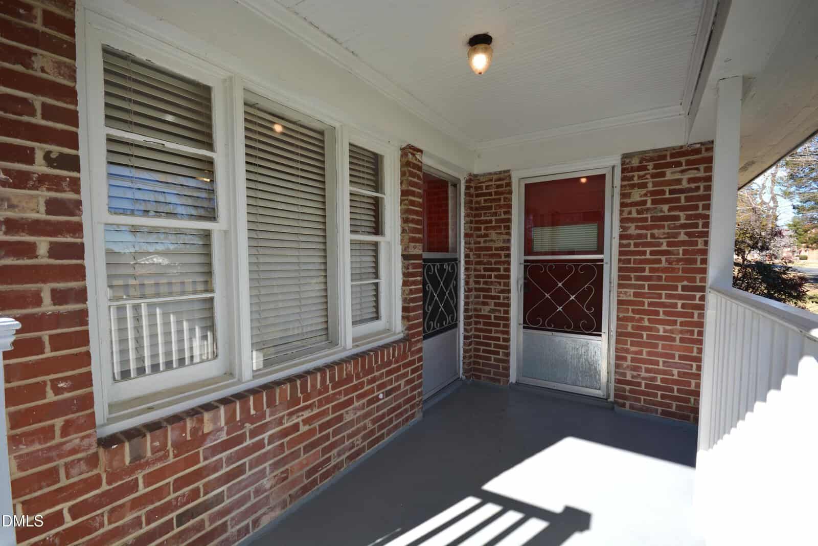 A covered front porch with red brick walls, a gray painted floor, white trim, three windows with blinds, a light fixture on the ceiling, and a red and silver screen door.