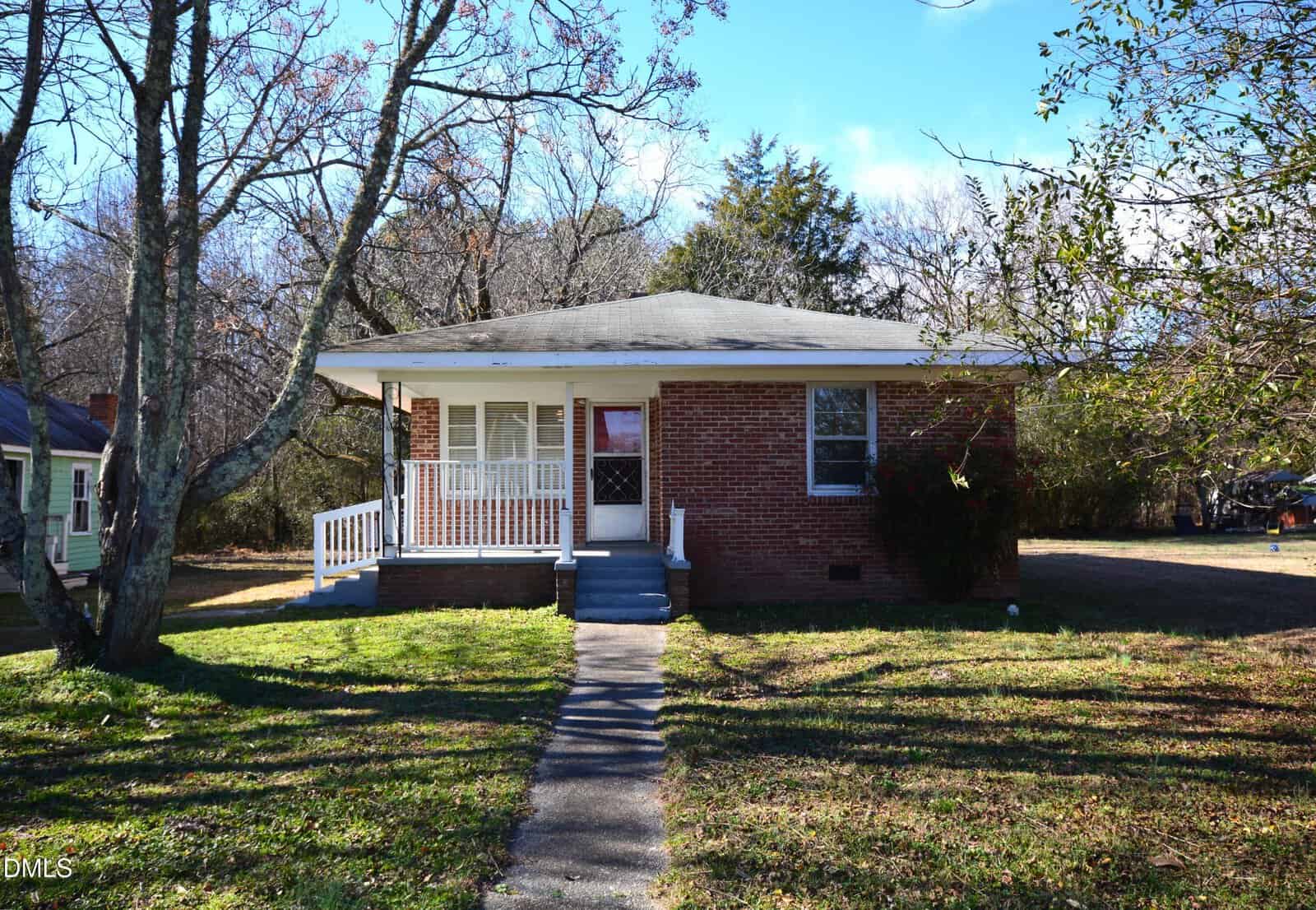 A small, single-story brick house at 1324 Raleigh Road in Oxford with a covered front porch, white railing, and a concrete walkway leading to the door. The yard has patchy grass, leafless trees, and some bushes under a clear blue sky.