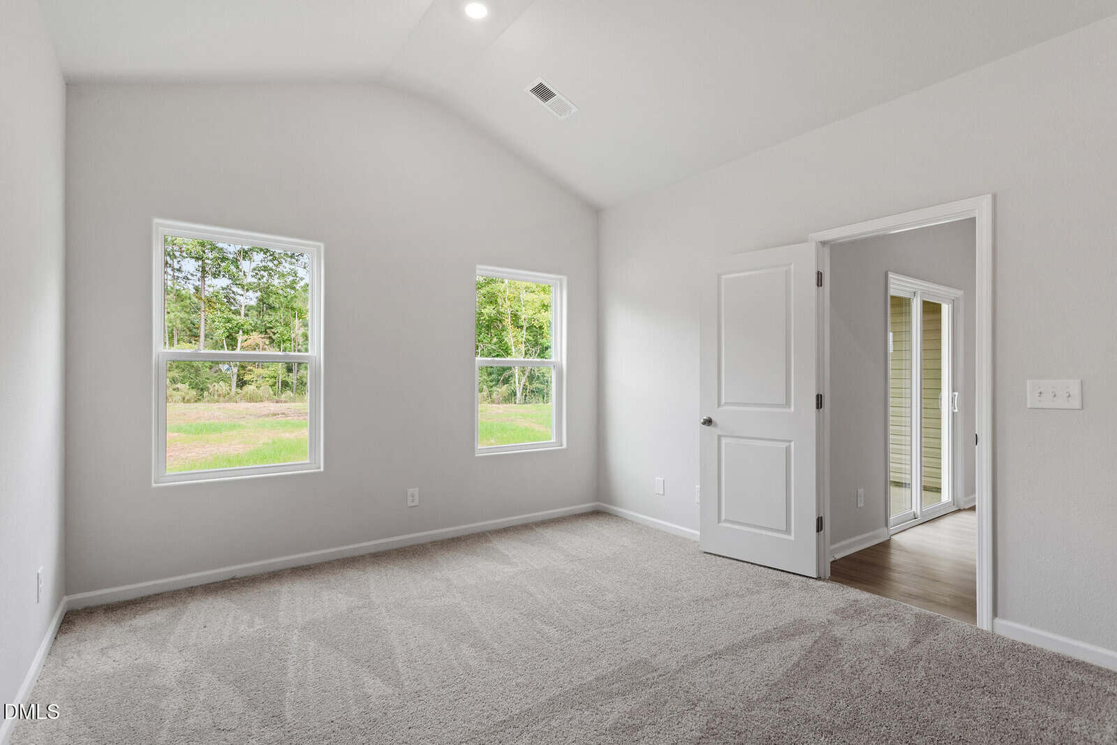 A bright, empty room with light-colored walls, two windows, and beige carpet. An open door leads to a hallway with wood flooring. The windows provide a view of green trees and grass outside.