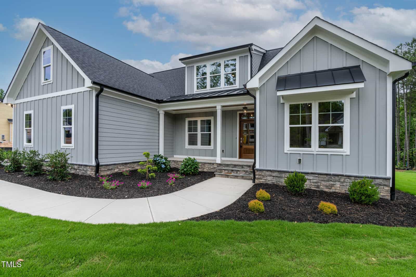 Single-story gray house with white trim, black roof, stone accents, and large front windows. A concrete walkway curves through a landscaped front yard with shrubs and a green lawn.
