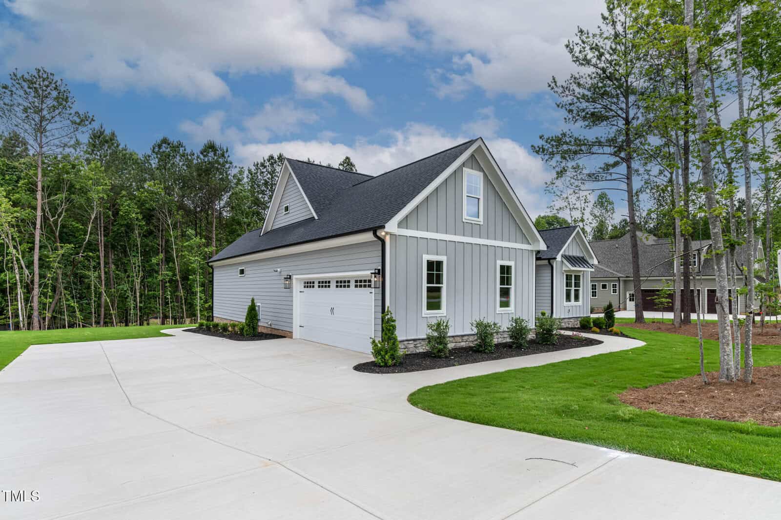 A light gray single-story house with white trim and a steep roof sits beside a large, curved concrete driveway. The yard is landscaped with green grass, small shrubs, and surrounded by tall trees.