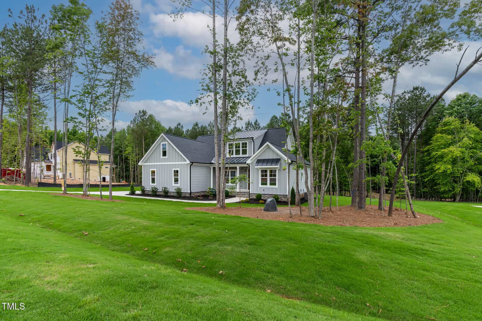 A modern, single-story gray house with white trim sits on a large, manicured lawn surrounded by tall trees. The sky is partly cloudy, and another house is visible in the background to the left.