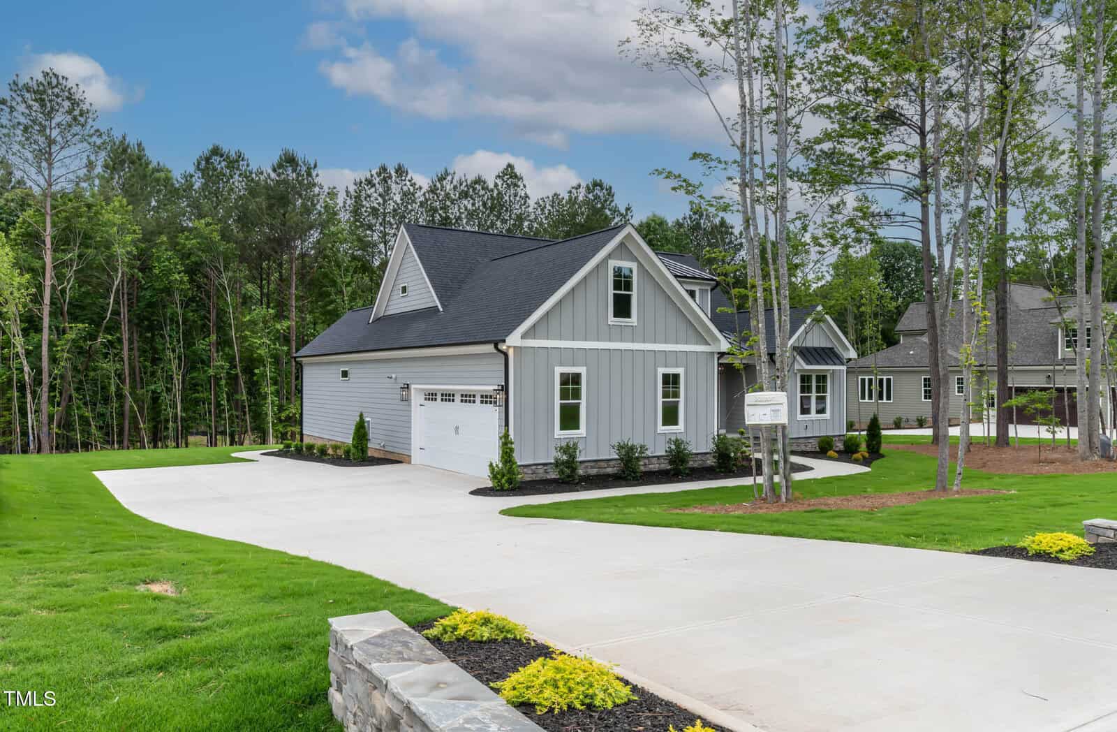 A modern gray house with white trim, a two-car garage, and a long concrete driveway, surrounded by green grass, landscaped shrubs, and tall trees under a partly cloudy sky.