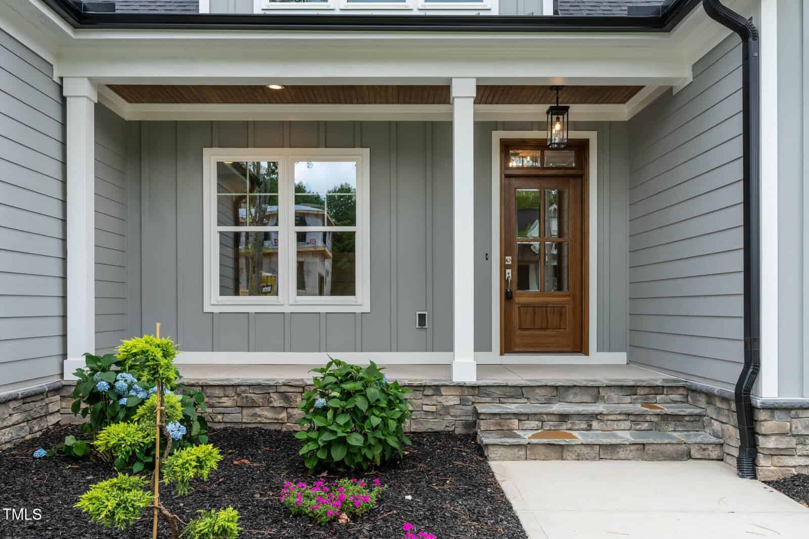 A modern house entryway with gray siding, a wooden front door with glass panels, white trim, a stone porch, two columns, and a small landscaped garden with shrubs and flowers.