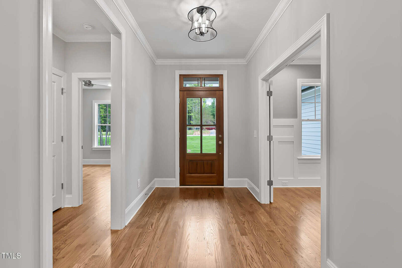 A hallway with light gray walls, wooden floors, and a wooden front door with glass panels; rooms with windows and doors are visible on both sides, and a ceiling light fixture is above.