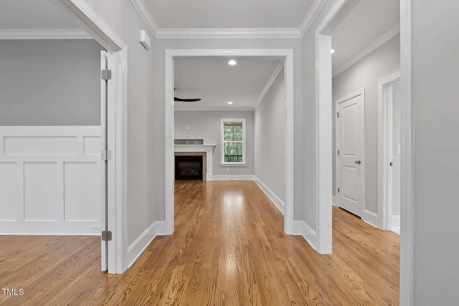 A hallway with polished wood floors, light gray walls, white trim, and several doorways leading to different rooms. A fireplace and large window are visible in the background living area. Ceiling lights are turned on.