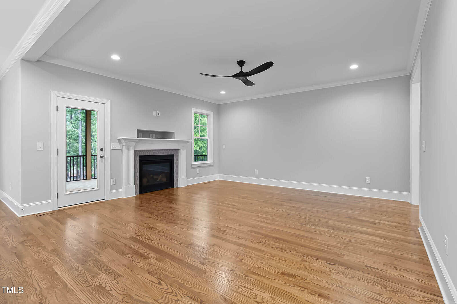 A spacious, empty living room with light gray walls, hardwood floors, a ceiling fan, a fireplace with a mantel, recessed lighting, a window, and a glass door leading to an outdoor area with greenery visible.