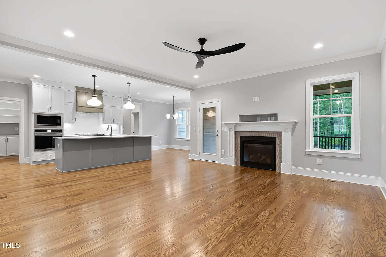 Spacious open-concept living area with light hardwood floors, a modern ceiling fan, a white fireplace, large windows, and a kitchen with an island, pendant lights, and white cabinetry in the background.