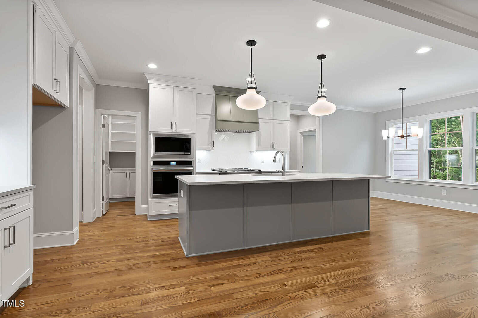 Modern kitchen with white cabinets, stainless steel appliances, a large gray island with a sink, hardwood floors, pendant lights, and an adjoining dining area with a window.