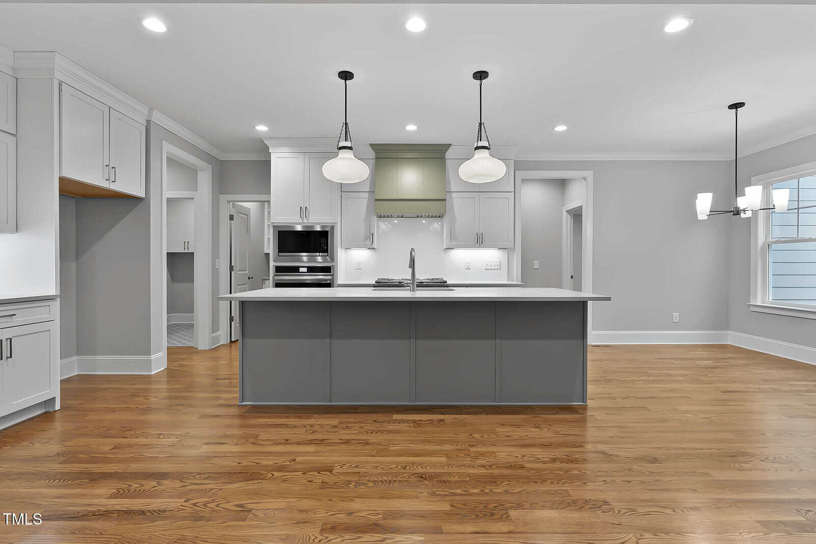 Modern kitchen with gray island, white cabinets, stainless steel appliances, two pendant lights, and hardwood floors. The space is brightly lit with recessed lighting and natural light from a window on the right.