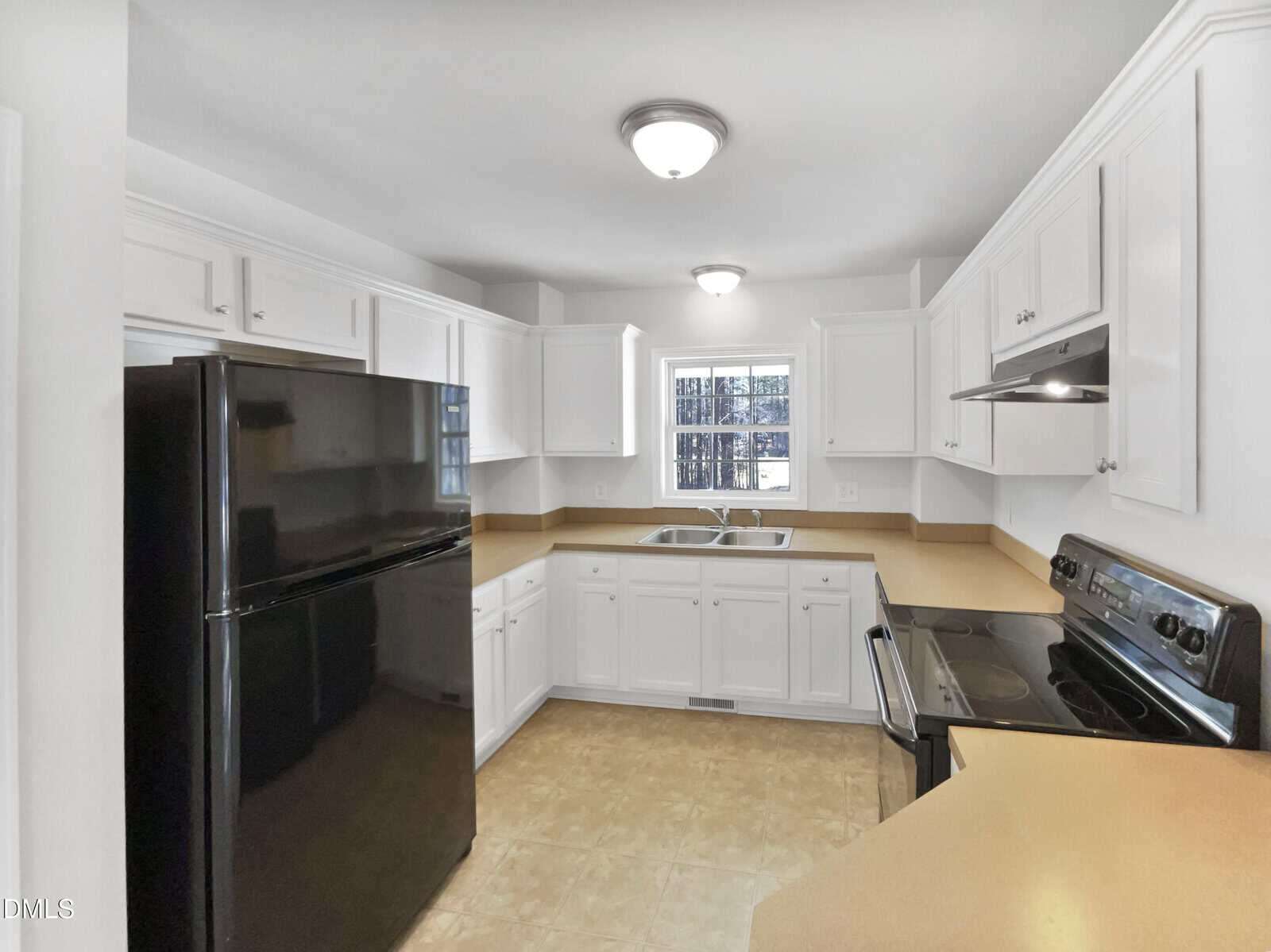 A kitchen with white cabinets, black appliances including a refrigerator and stove, beige countertops, a double sink under a window, and light-colored tile flooring. Ceiling lights illuminate the space.