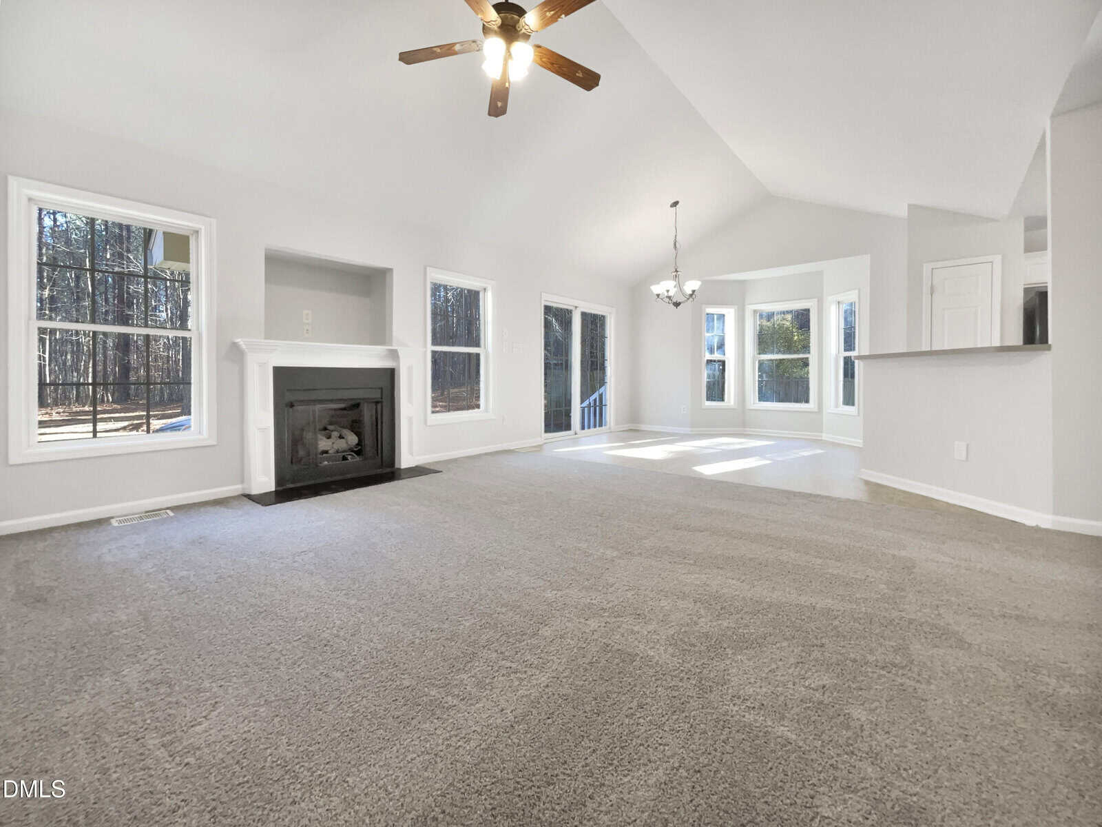 A spacious, empty living room with light gray carpet, white walls, a ceiling fan with lights, a fireplace, large windows, and an adjacent dining area with a bay window and overhead chandelier.