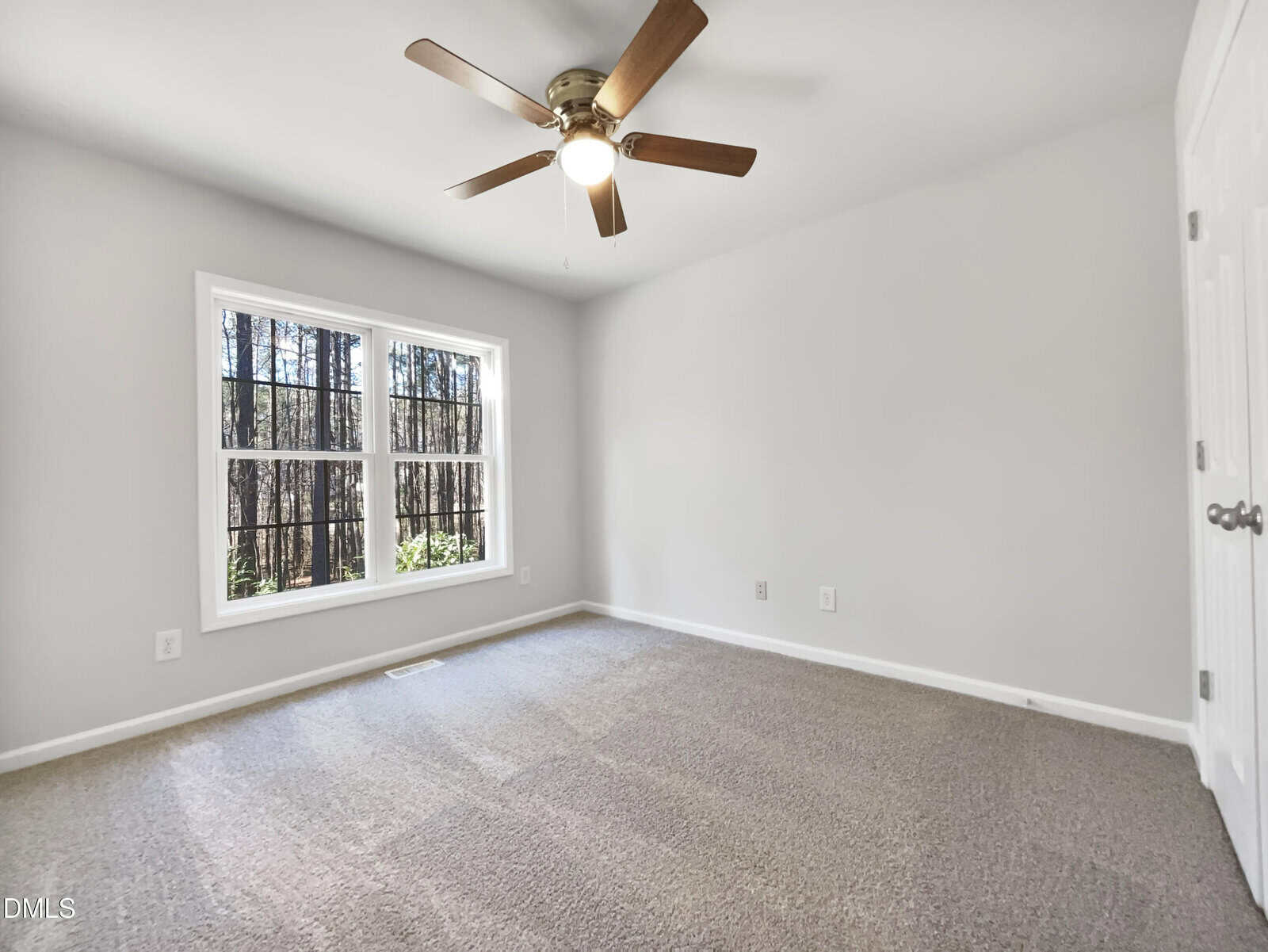 A vacant room with beige carpet, light gray walls, a ceiling fan with light, and a window with black trim providing a view of trees outside. Two closed white doors are visible on the right.