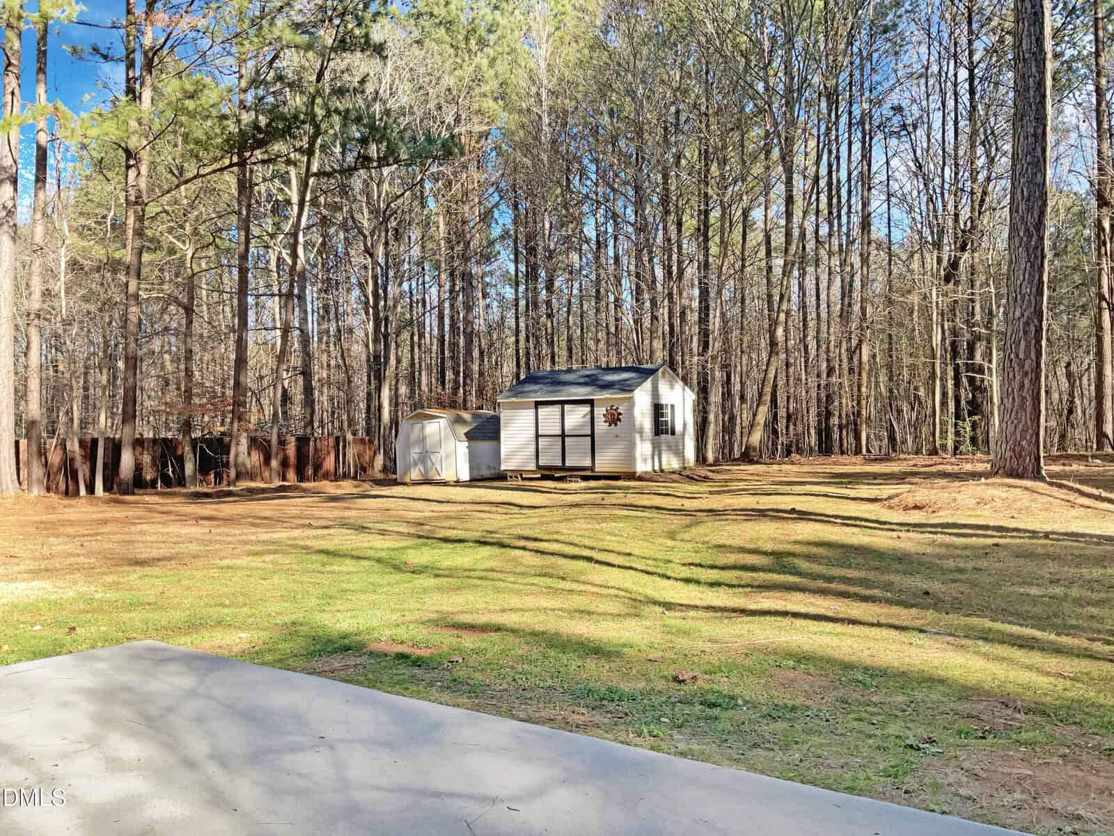 A small white shed with a dark roof and a decorative star stands in a grassy, partially shaded yard bordered by tall pine trees. A smaller structure is nearby, and part of a concrete slab is visible in the foreground.