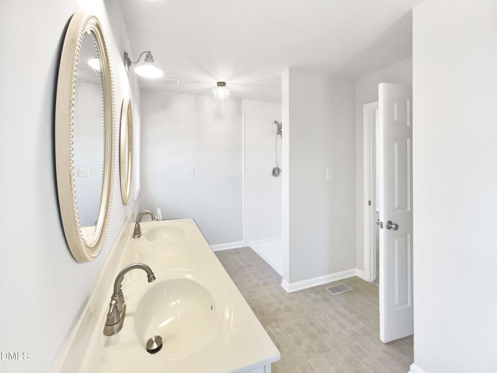 A bright bathroom with double sinks, two round mirrors, a walk-in shower, white walls, and beige floor tiles. The door is open, and a ceiling light fixture is visible.