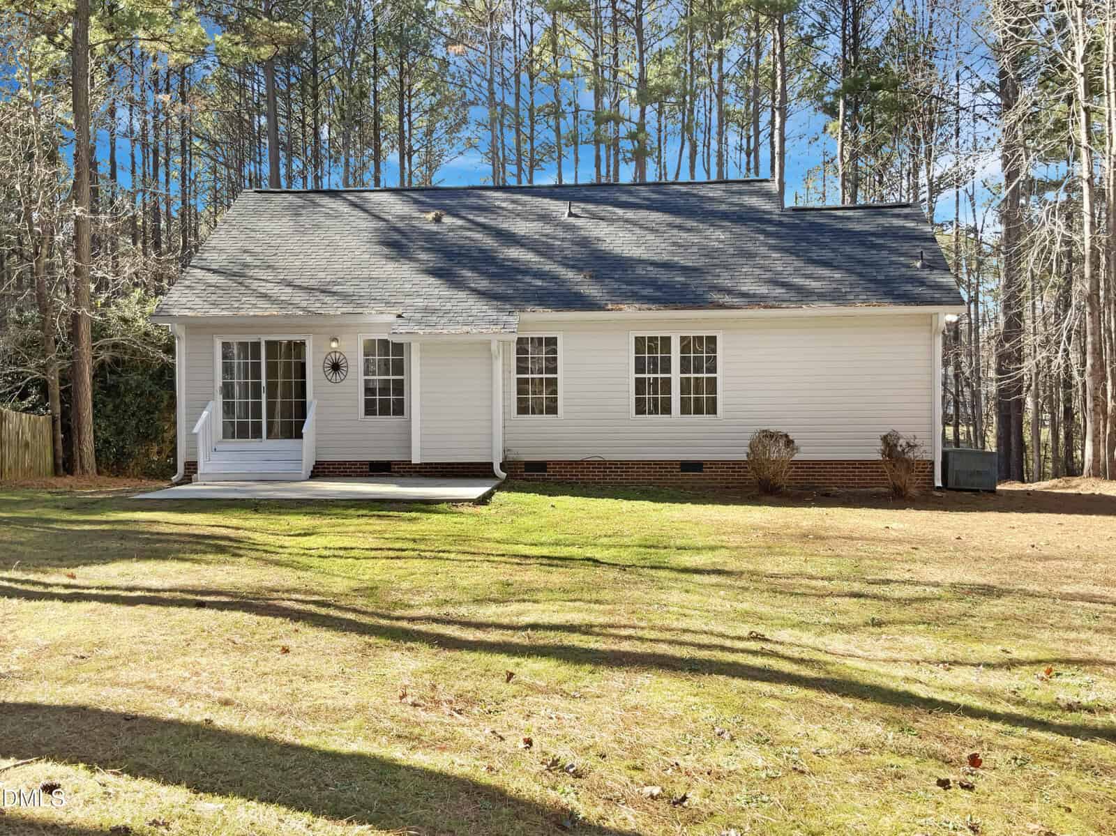 A single-story white house with a dark shingled roof, three large windows, and a small porch with steps. The house sits on a grassy yard bordered by tall pine trees in the background.