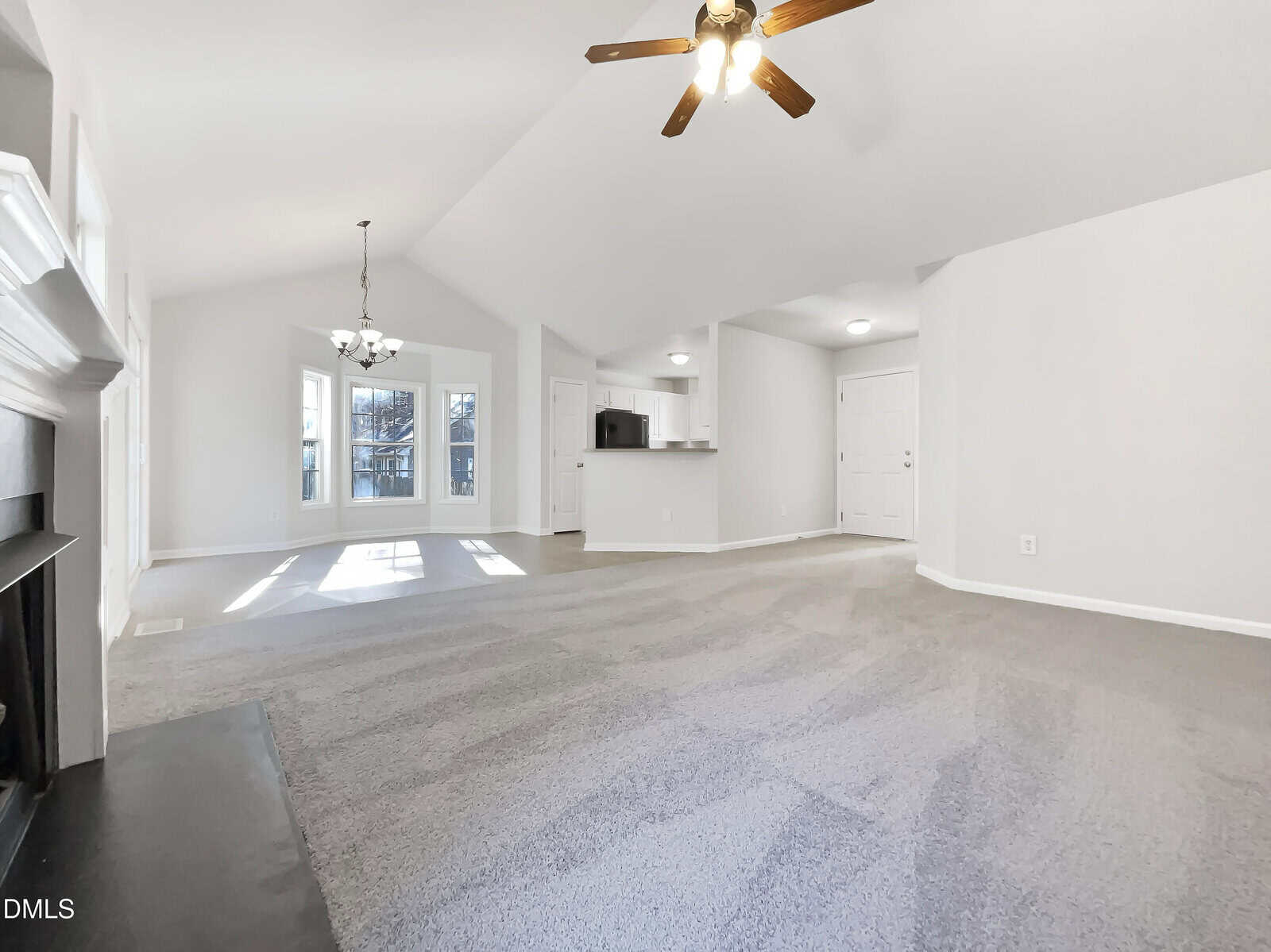 Spacious living area with gray carpet, white walls, a ceiling fan, fireplace on the left, dining area near large windows, and an open kitchen with a breakfast bar. White door and entryway are visible in the background.