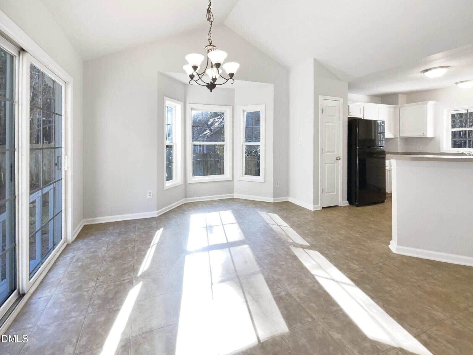 A bright, empty room with large windows, a hanging light fixture, tiled flooring, and an open view into a kitchen with white cabinets and black appliances. Sunlight streams across the floor.
