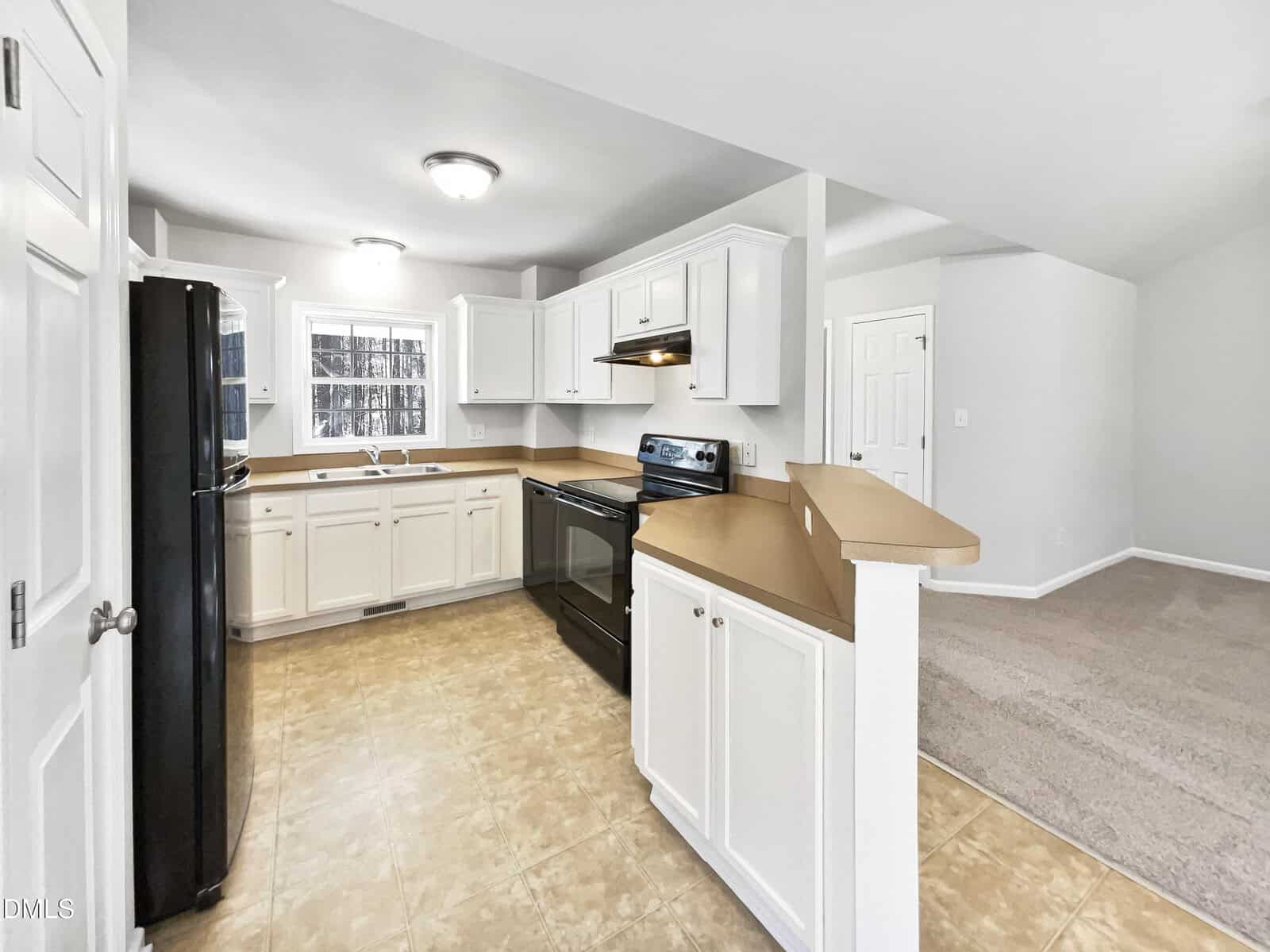 A kitchen with white cabinets, beige countertops, a stainless steel stove, black refrigerator, and tiled flooring. The space is open to a carpeted living area and has neutral-colored walls.