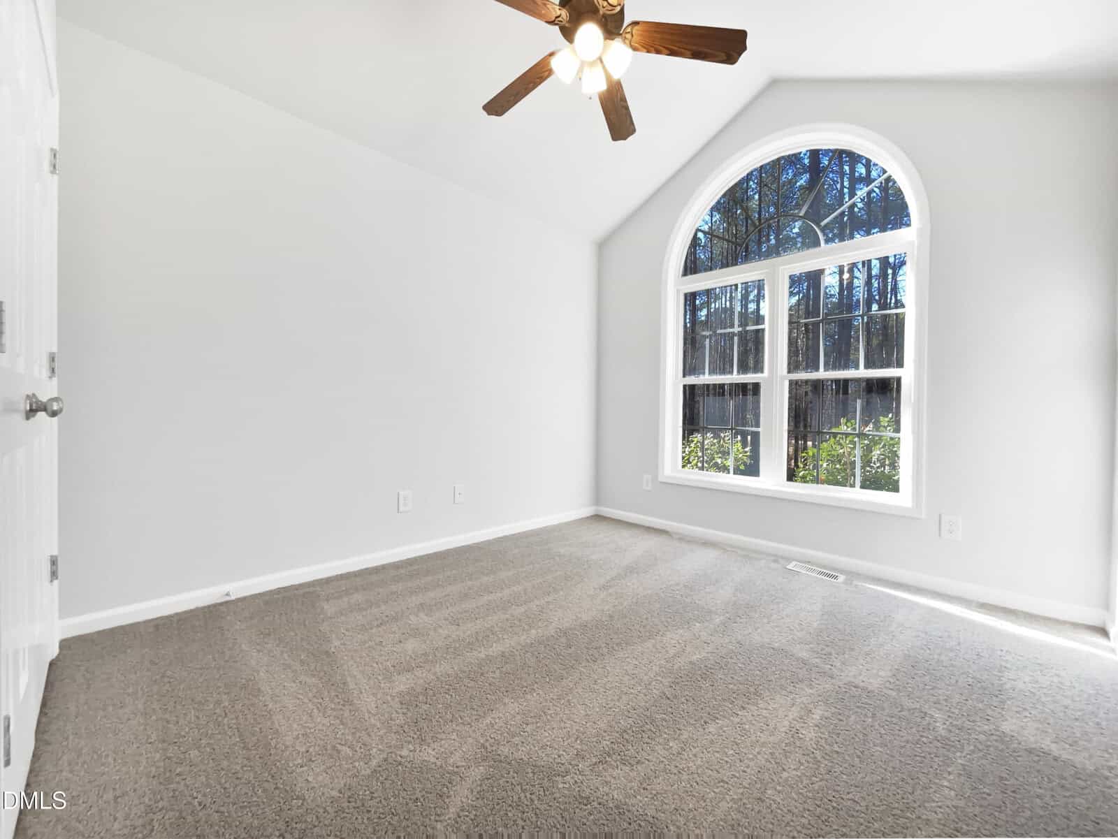 Bright, empty room with light gray walls, carpeted floor, and a large arched window showing trees outside. A ceiling fan with lights hangs from the vaulted ceiling.