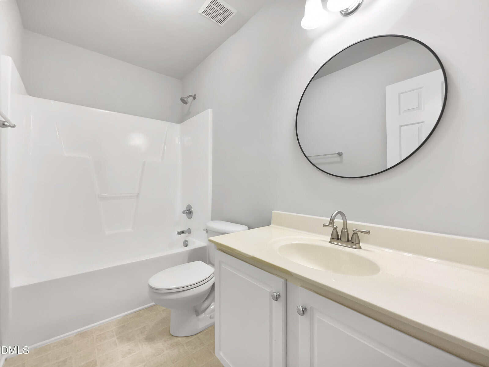 White bathroom with a bathtub and shower, toilet, single sink vanity, oval mirror, and overhead light. The walls and cabinets are white, and the countertop is beige. The floor is light-colored tile.