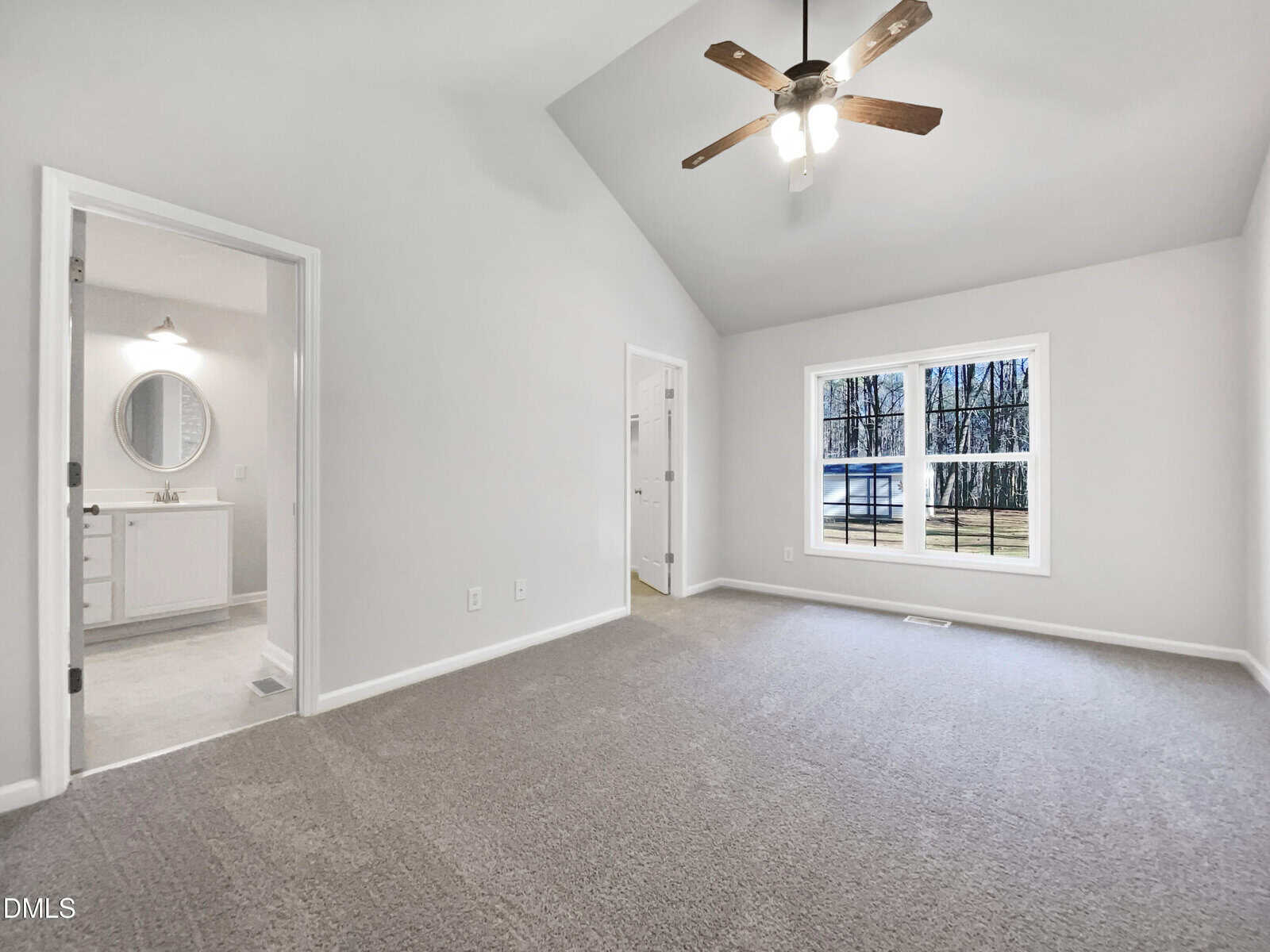 Bright, empty bedroom with beige carpet, white walls, ceiling fan, large window showing trees, and two doorways leading to a bathroom and a closet.