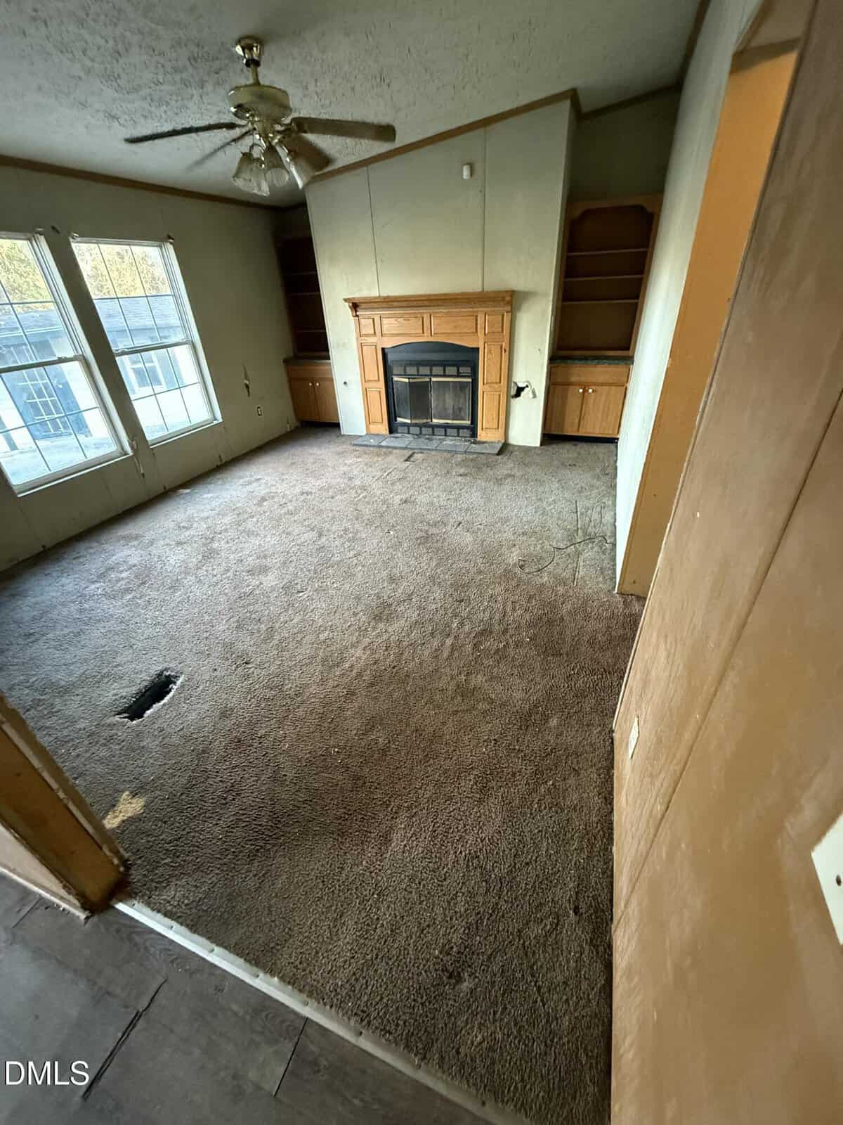 A living room with brown carpet, a ceiling fan, a fireplace centered between built-in shelves, two large windows, and a visible hole in the carpet near the wall. The room appears worn and needs maintenance.