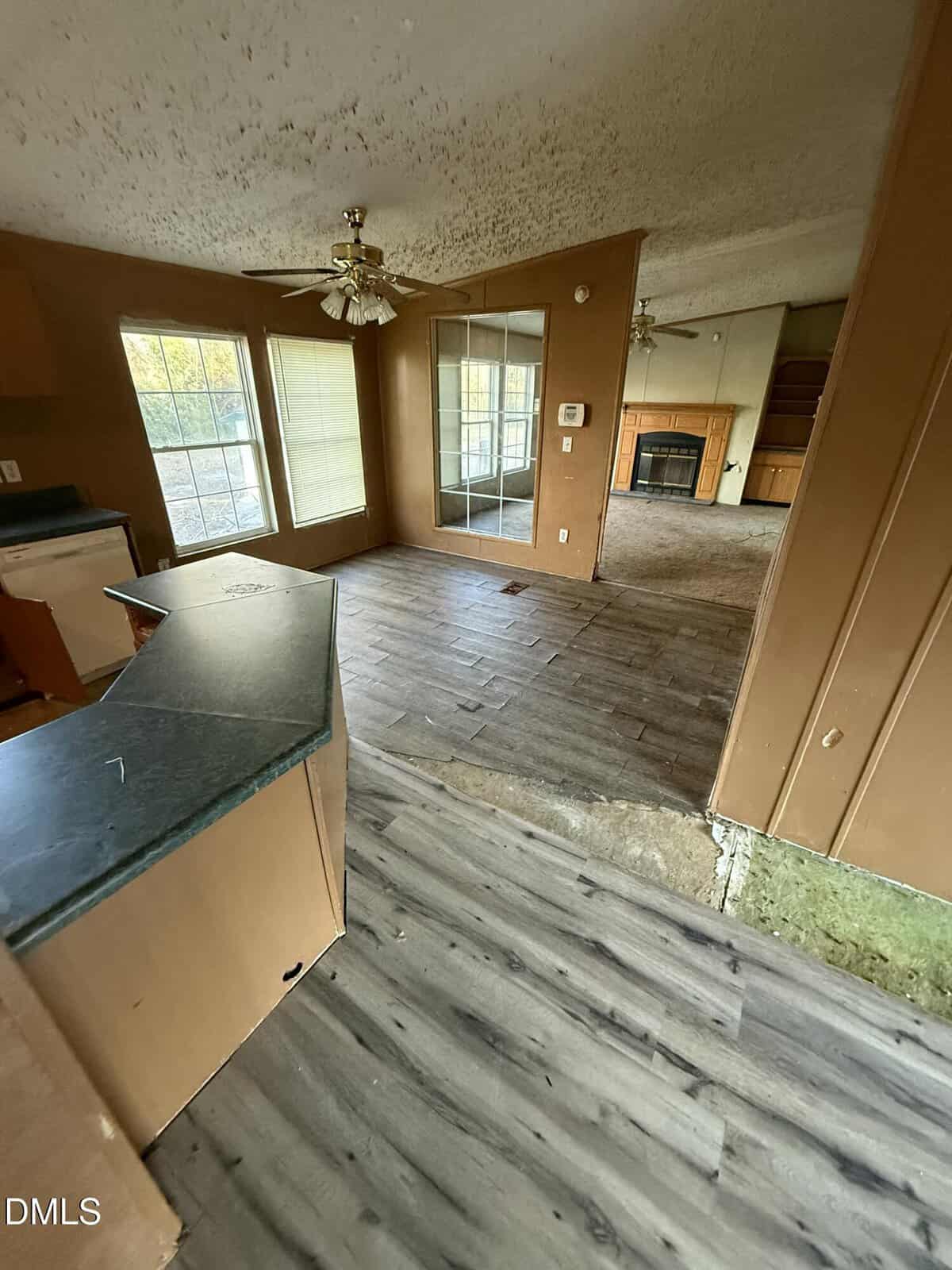 A view of an empty room with brown walls and wood-look flooring, showing a kitchen counter, large windows, a ceiling fan, a mirrored wall, and a fireplace in the background. Some flooring appears unfinished.