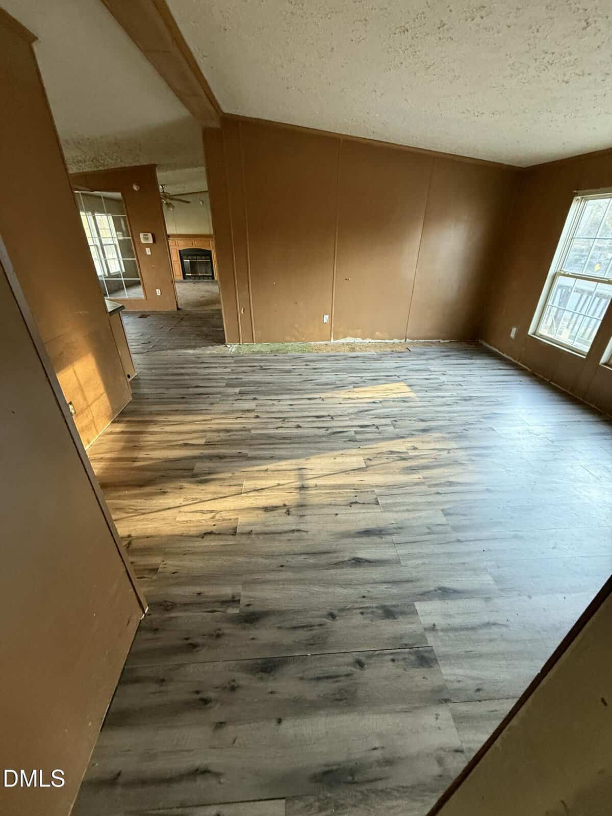 Room with brown walls and light wood-patterned vinyl flooring, viewed from a doorway. Two large windows on the right let in natural light. An adjoining room with matching walls and floor is visible in the background.