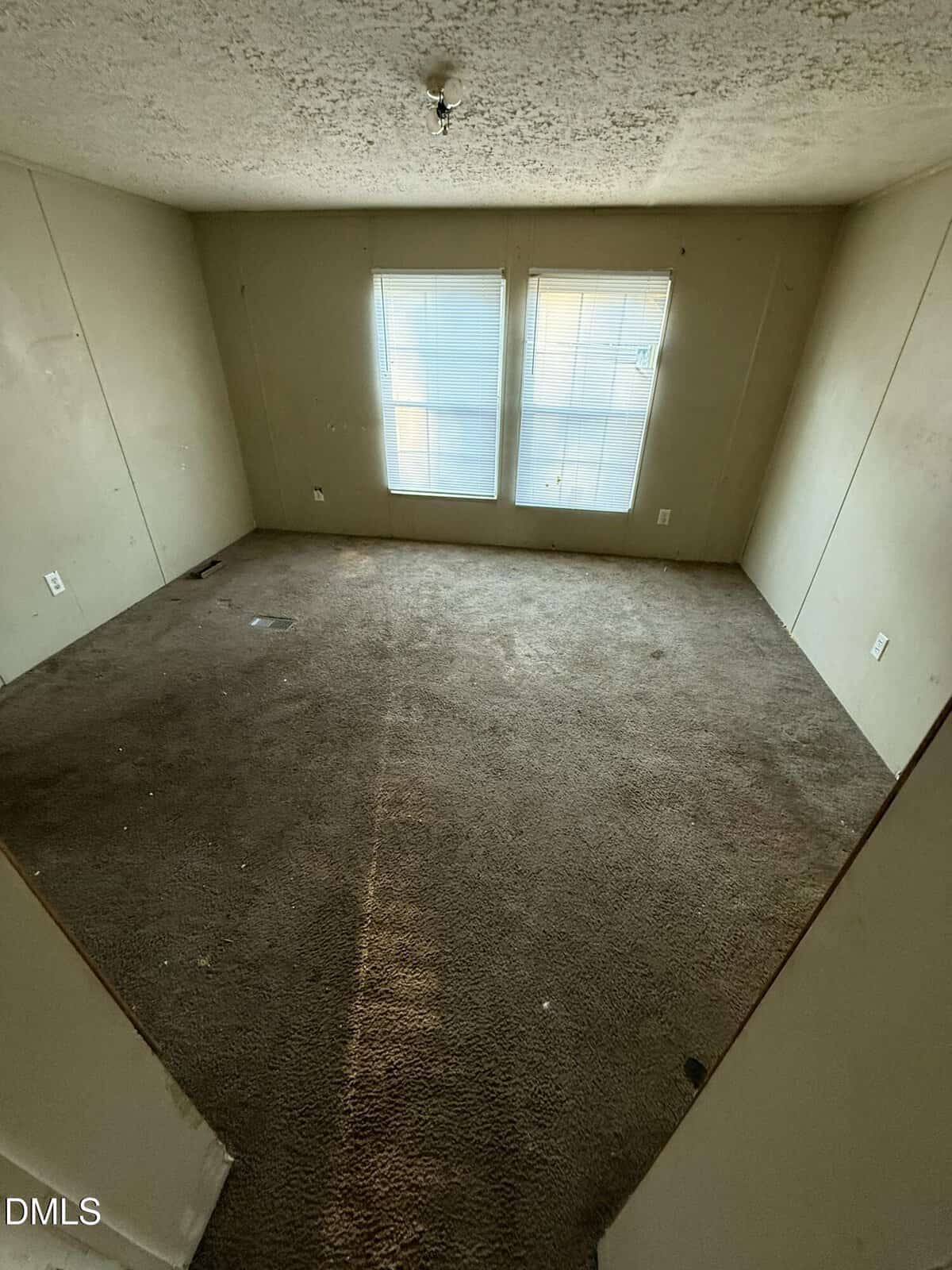 Empty room with beige walls, two large windows letting in light, and worn brown carpet. Ceiling has visible texture and an exposed light fixture. Walls and carpet show some signs of wear.