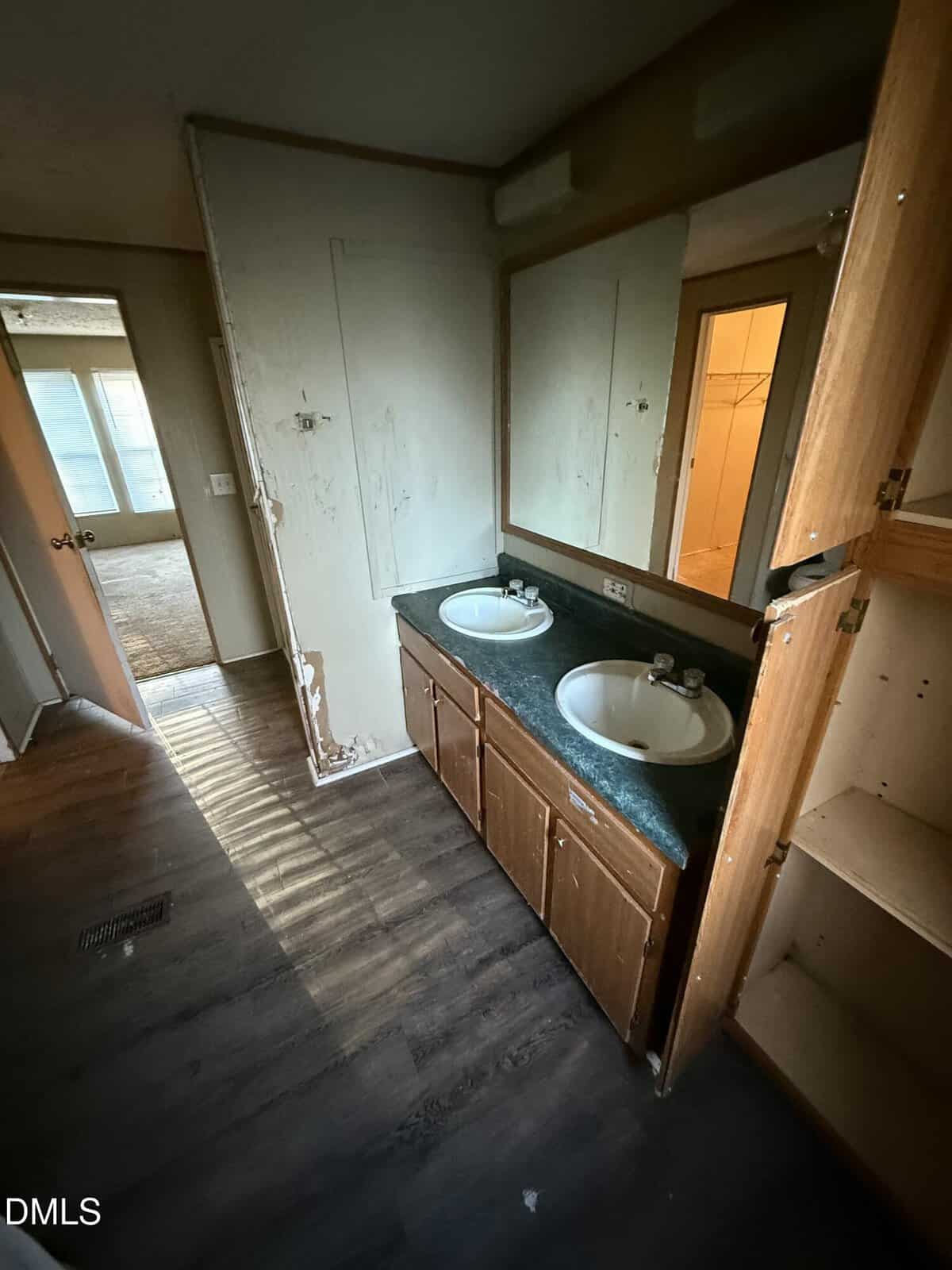 A bathroom with a double sink vanity, dark countertop, wood cabinets, and a large mirror. The wall shows peeling paint, and some shelves are empty. Light from a nearby room shines through an open doorway.