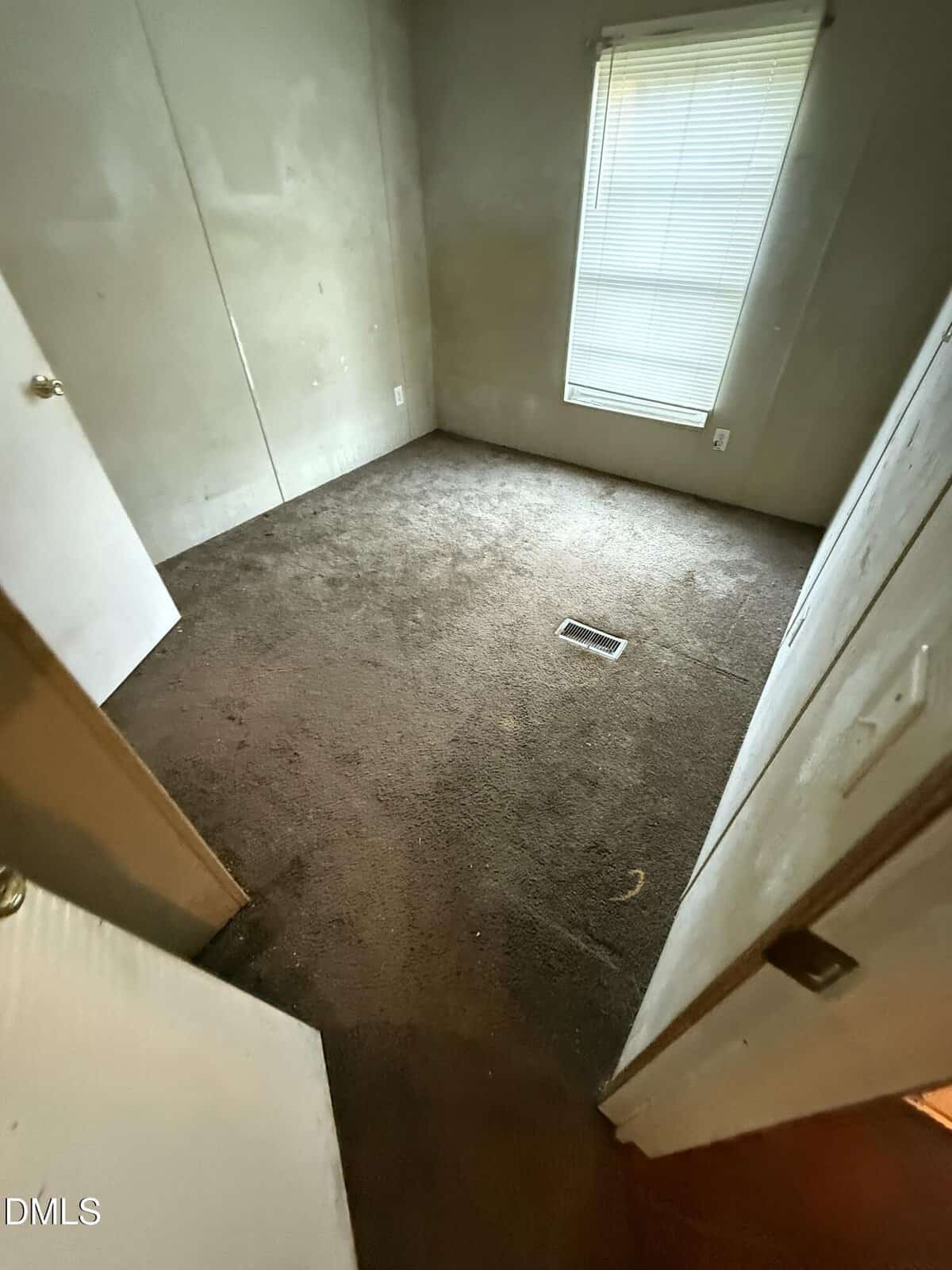 Small empty room with beige walls, brown carpet, a closed window with blinds, a floor vent in the center, and an open door leading into the space. The lighting is dim, and the room appears unoccupied.