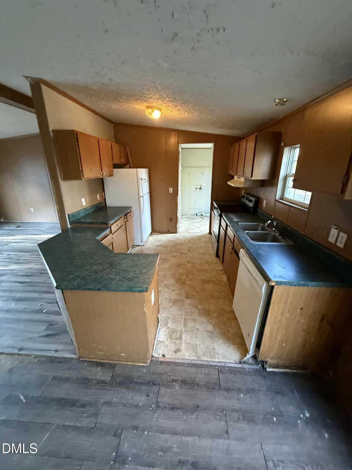 A small, outdated kitchen with wood cabinets, green counters, white appliances, and a beige linoleum floor. The area has visible wear, and the adjacent room has dark wood flooring.