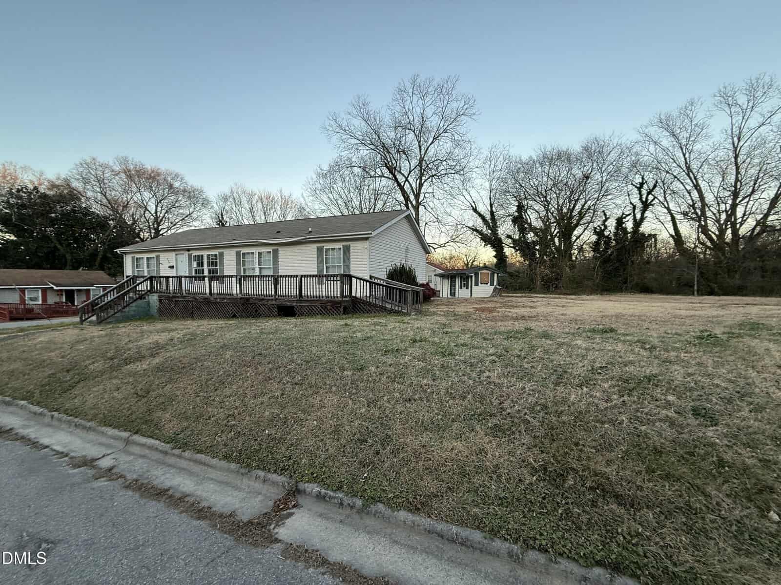 A single-story house with a large wooden ramp and deck sits on a slightly sloped grassy lot. Leafless trees and a small shed are in the background under a clear sky. A paved road runs in front of the property.