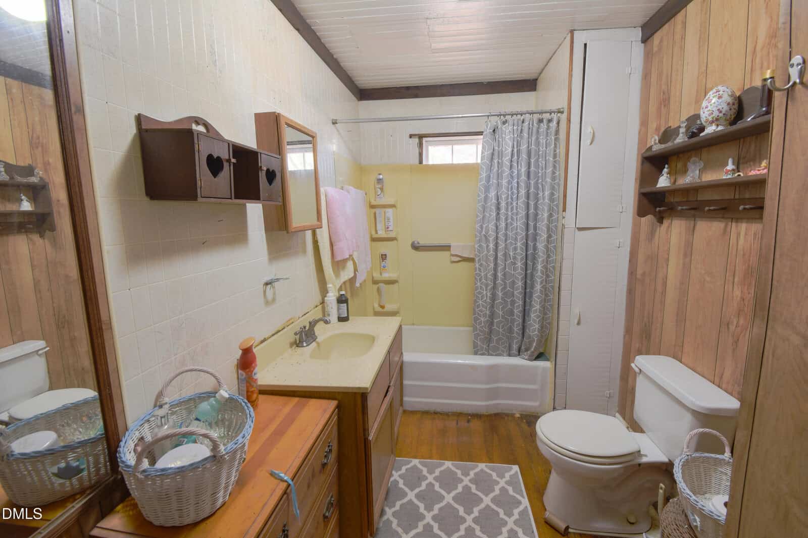 Small bathroom with wood-paneled walls, a bathtub with a shower curtain, a toilet, a sink with a mirror, shelves with decorations, towels, and a wicker basket on the floor. Light from a window above the tub.