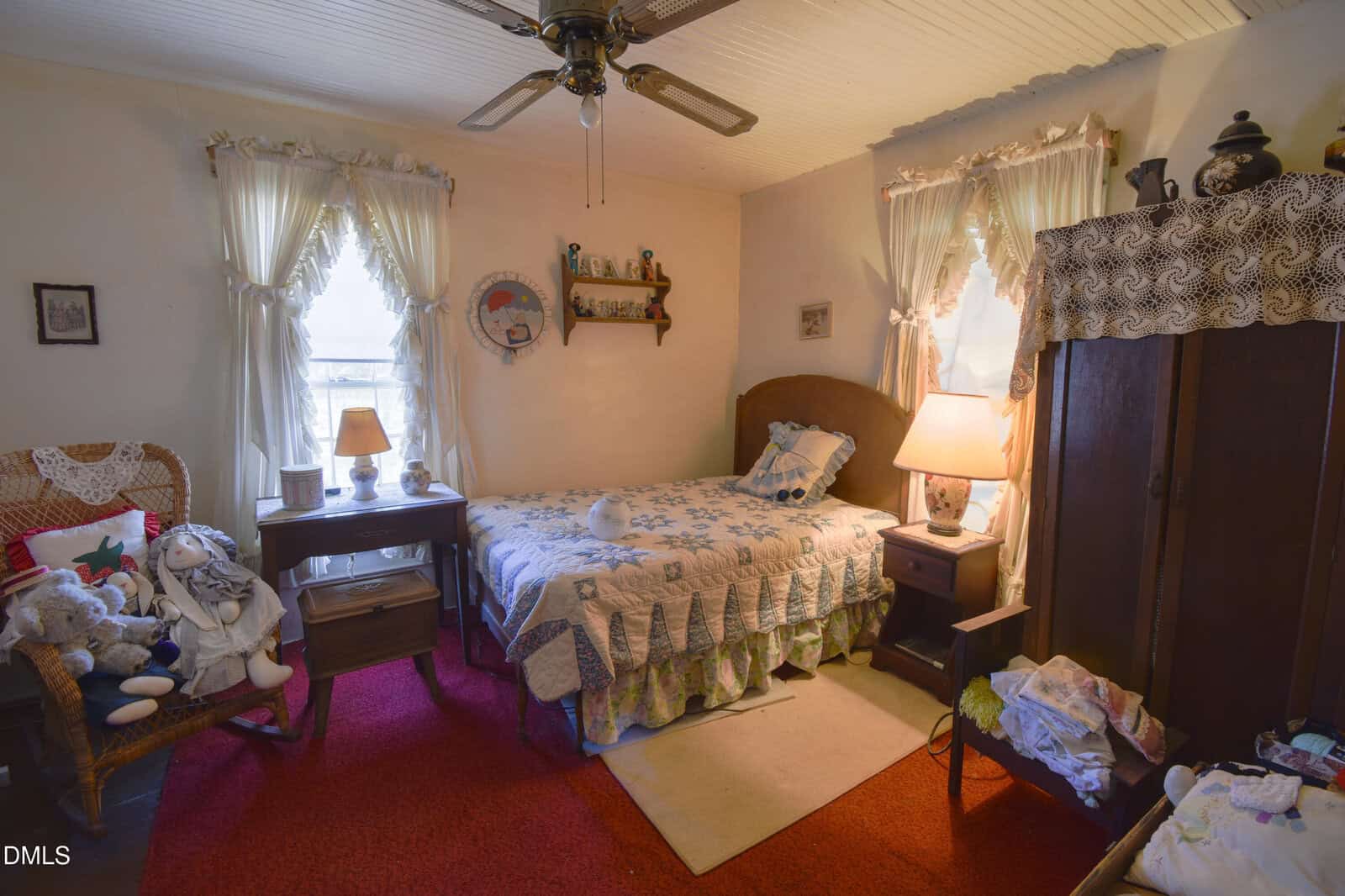 A cozy bedroom with a single bed, patchwork quilt, side tables with lamps, wicker chair with stuffed animals, red carpet, curtains on two windows, and various knick-knacks on shelves and furniture. Warm, inviting atmosphere.