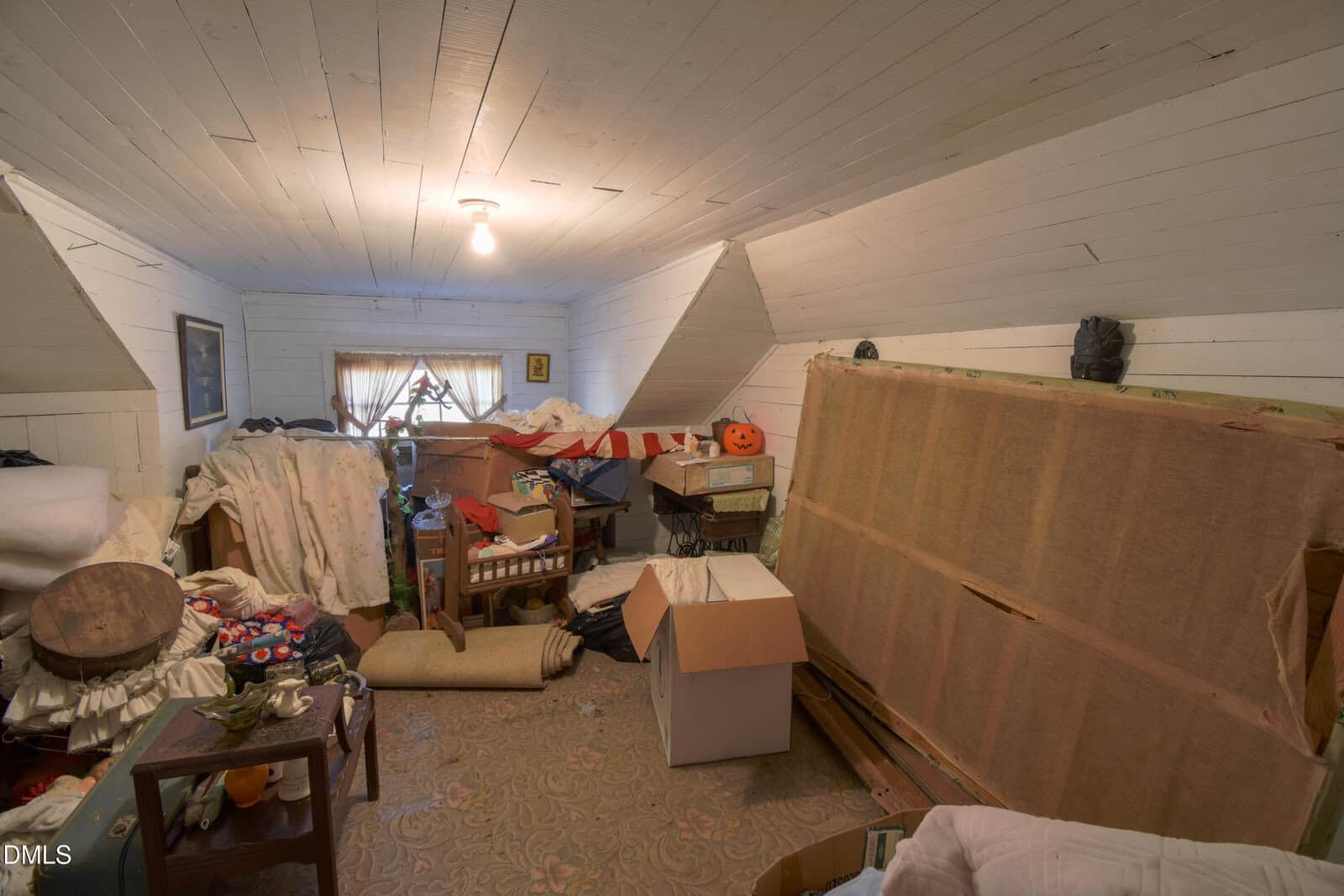 Cluttered attic room with slanted white ceiling and walls, scattered boxes, furniture, fabrics, and a window with sheer curtains at the far end.