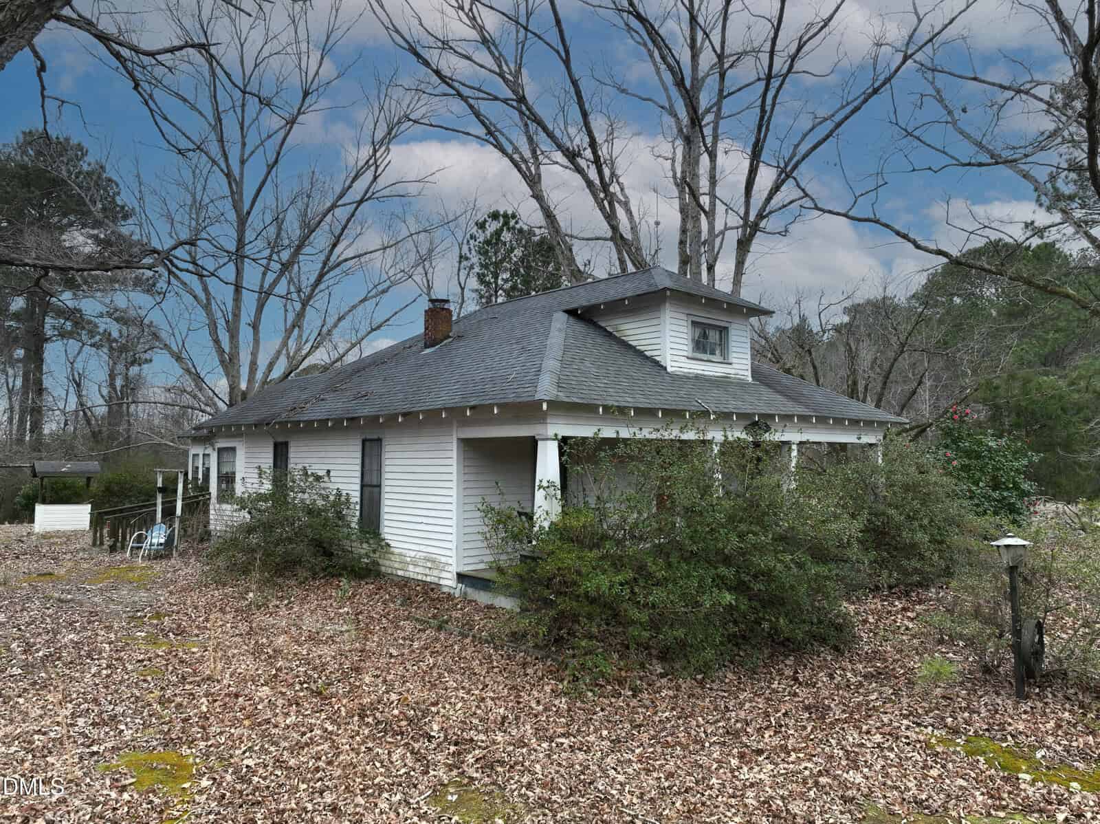 A small white house with a front porch sits among overgrown bushes and fallen leaves. Bare trees surround the property under a partly cloudy sky, creating a rustic, secluded atmosphere.
