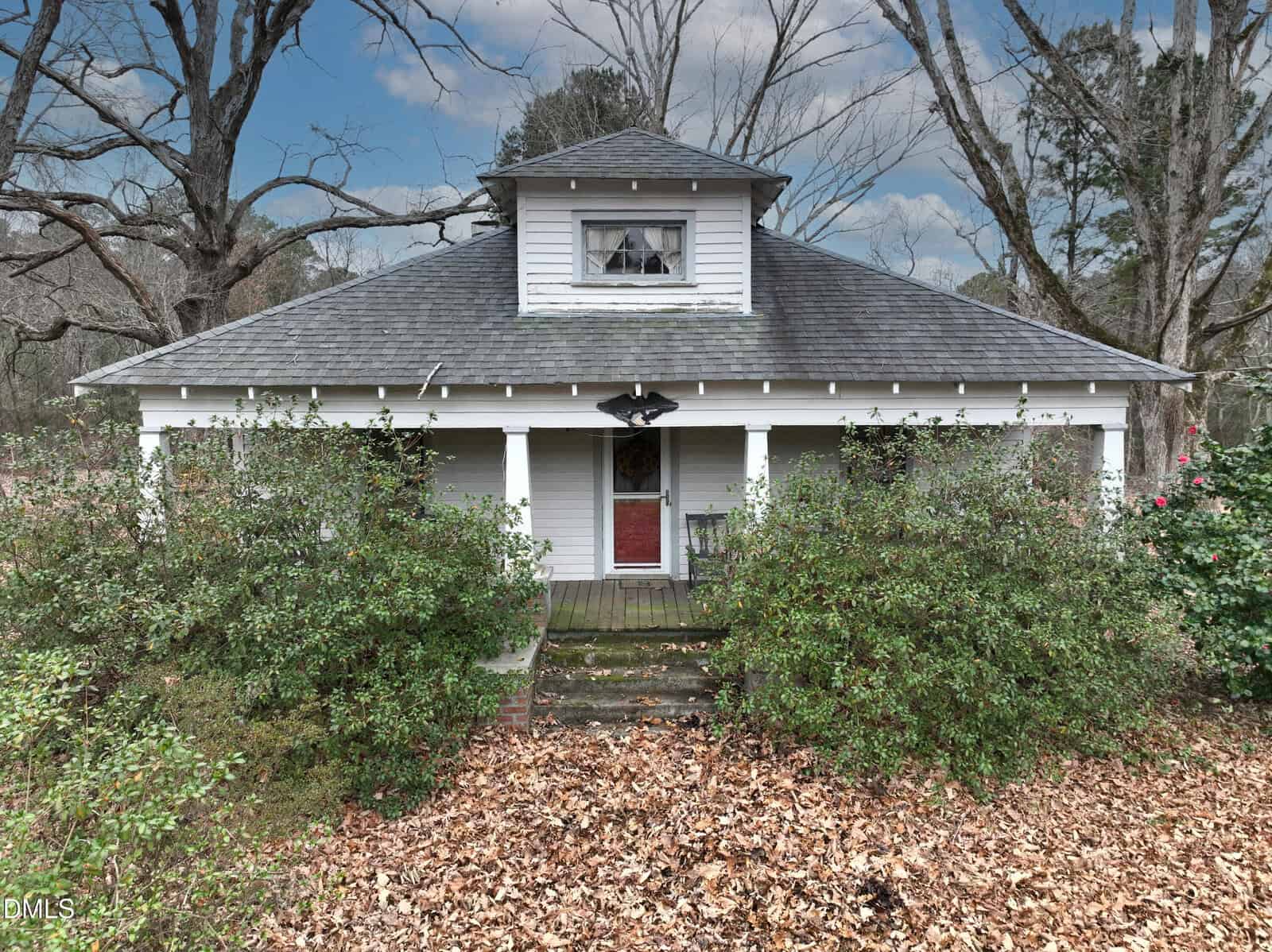 A small, white, single-story house at 847 Brantleytown Road in Zebulon features a central dormer window, a covered front porch with a red front door, overgrown bushes, fallen leaves, and leafless trees under a partly cloudy sky.
